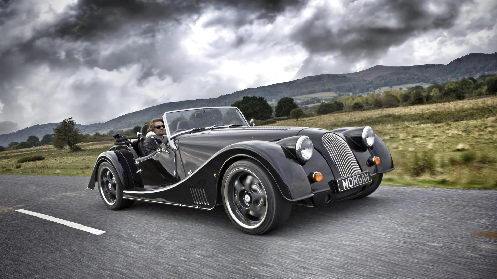 Black Morgan convertible sports car with chrome grille and round headlights driving on a road with cloudy sky and hills in the background.