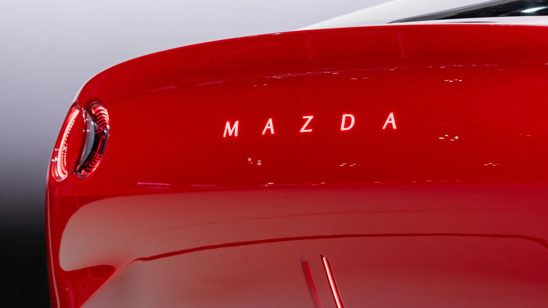 Glossy red rear of a Mazda car with illuminated "MAZDA" lettering and a circular taillight on the left.