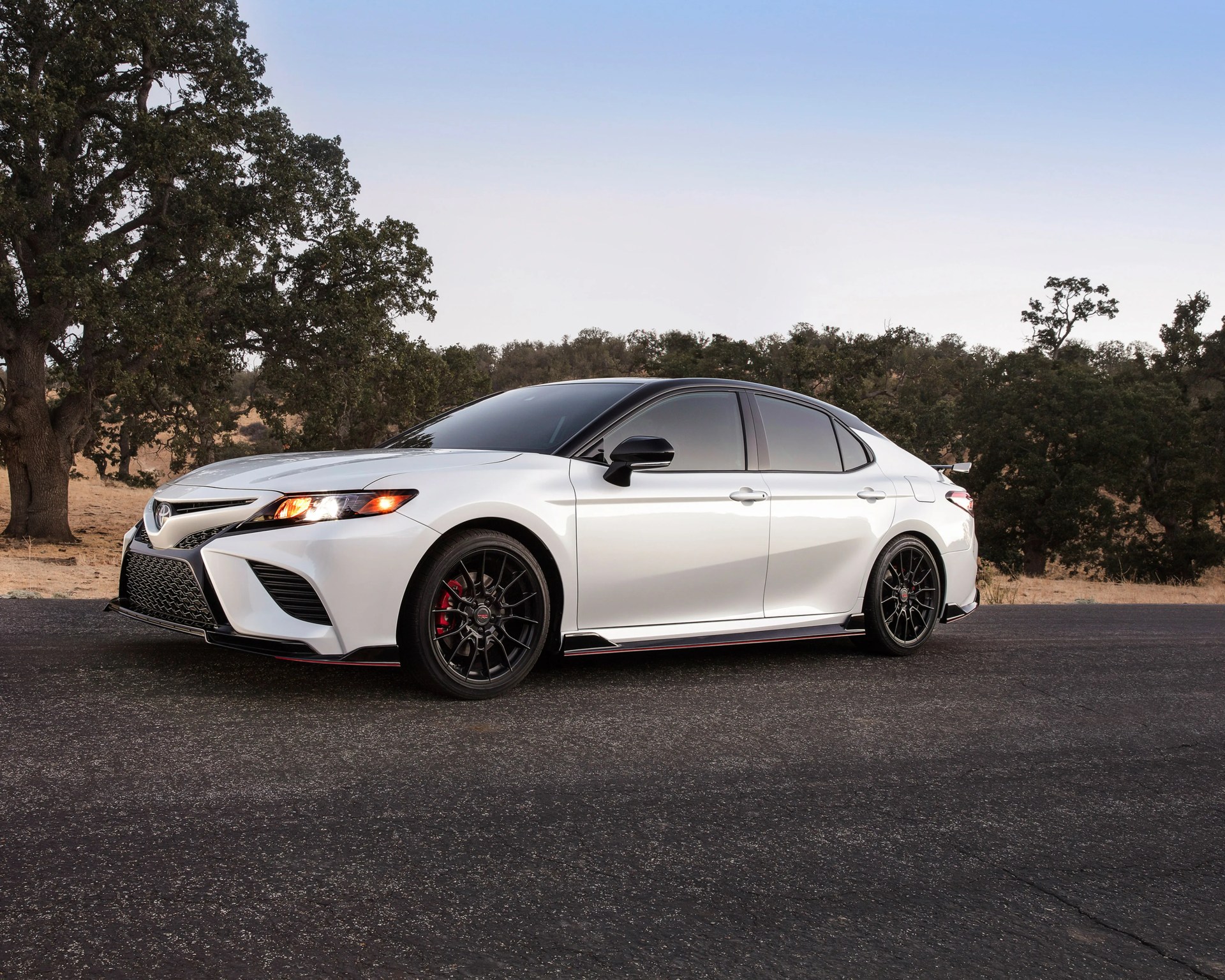 White Toyota sedan with black roof, black wheels, and red brake calipers parked on asphalt near trees.