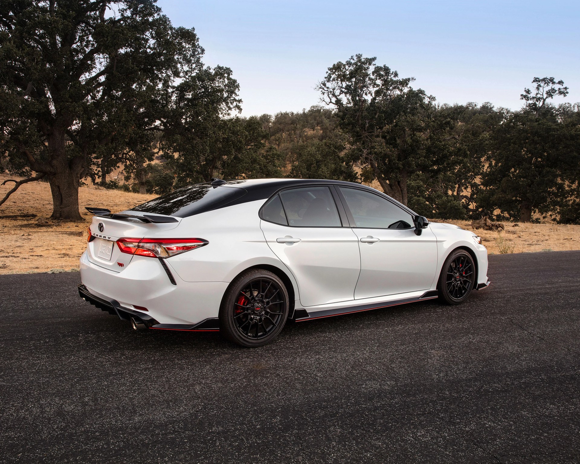 White Toyota Camry TRD sedan with black roof and black wheels parked on asphalt road near trees.