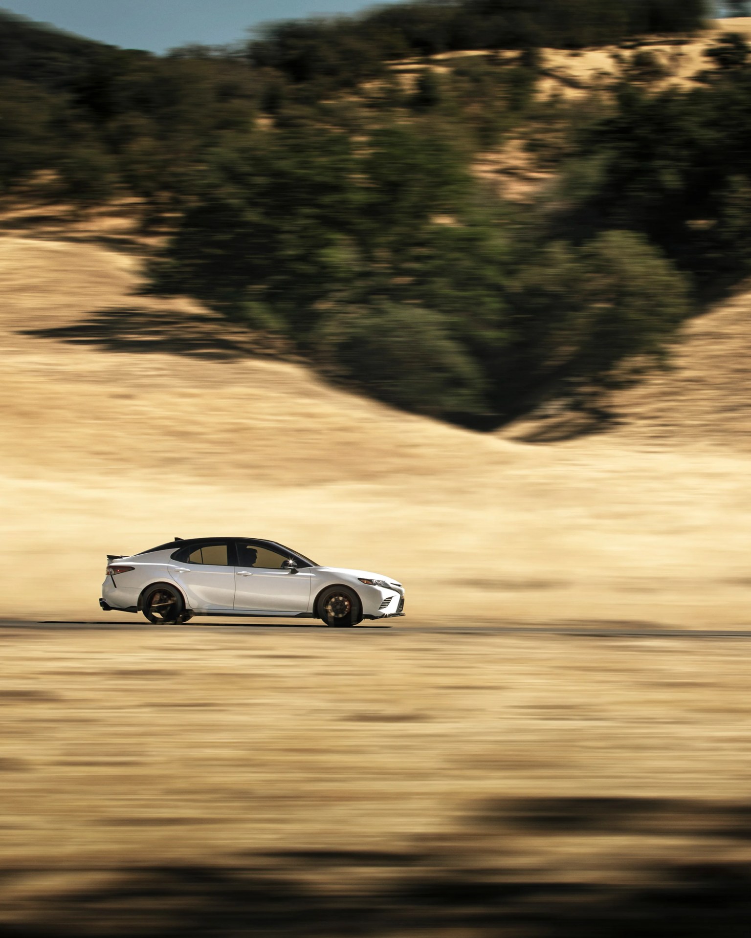 Silver sedan with black roof and tinted windows driving on a desert road with blurred background.
