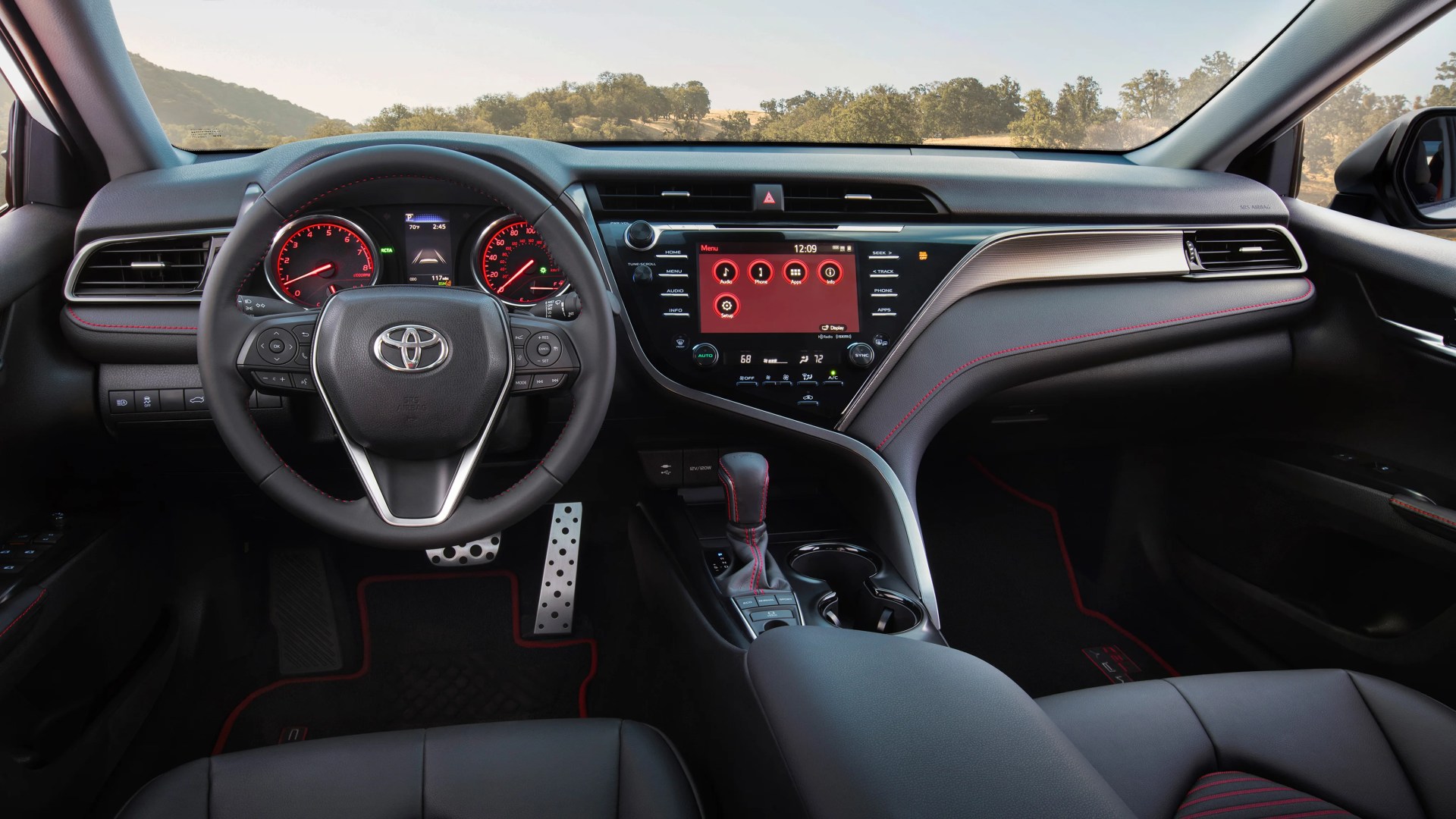 Black leather Toyota steering wheel with red stitching and digital dashboard, touchscreen center console, and gear shift in a car interior.
