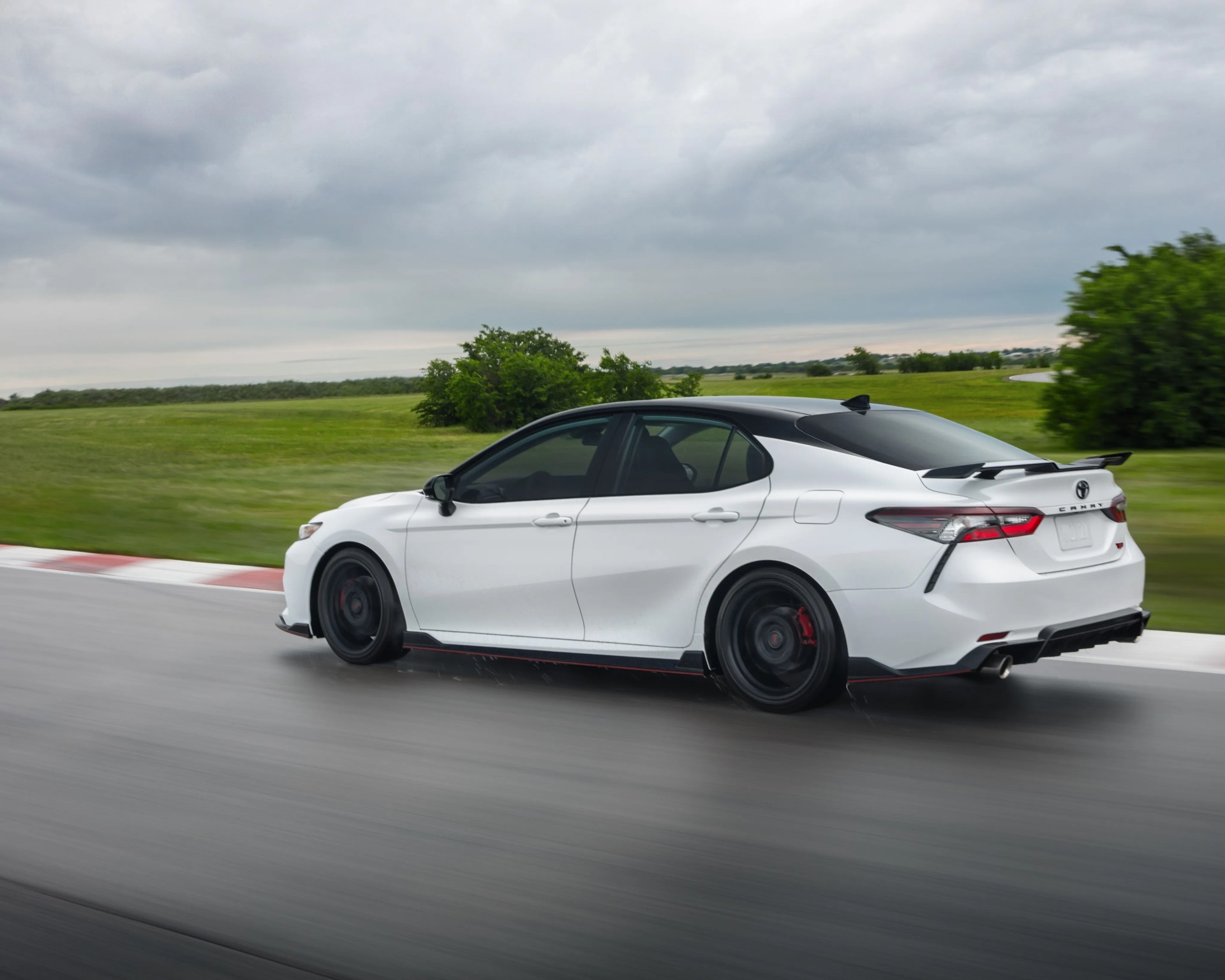 White Toyota Camry with black roof and rear spoiler driving on a wet racetrack with green fields and cloudy sky.