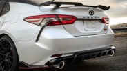 Rear view of a white Toyota Camry TRD with black spoiler, black wheels, and dual exhaust pipes on a road.