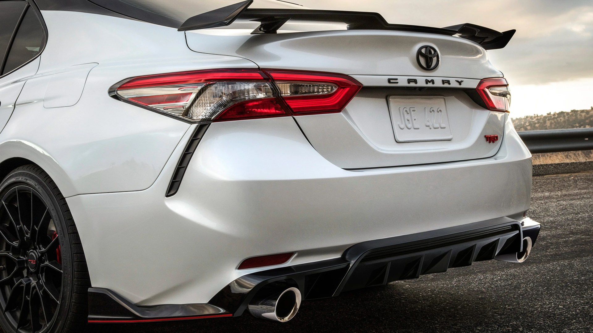 Rear view of a white Toyota Camry TRD with black spoiler, black wheels, and dual exhaust pipes on a road.