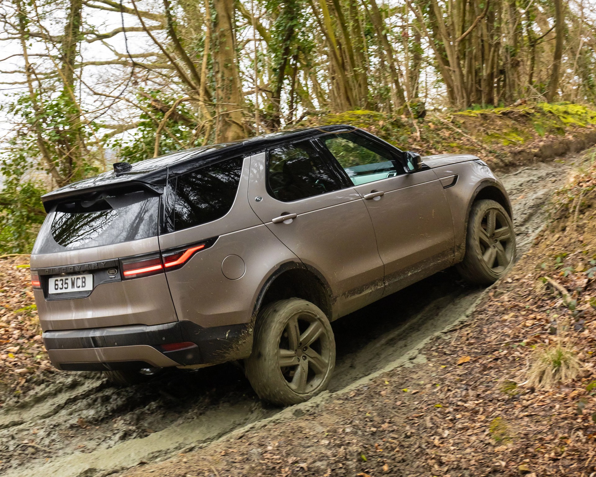 Brown Land Rover Discovery SUV with black roof driving off-road on a muddy forest trail.