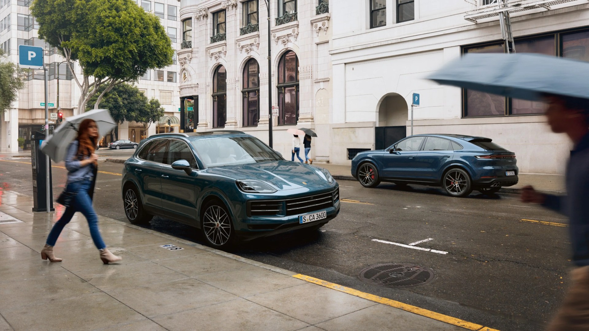 Two dark blue Porsche SUVs parked on a wet city street with people walking under umbrellas.