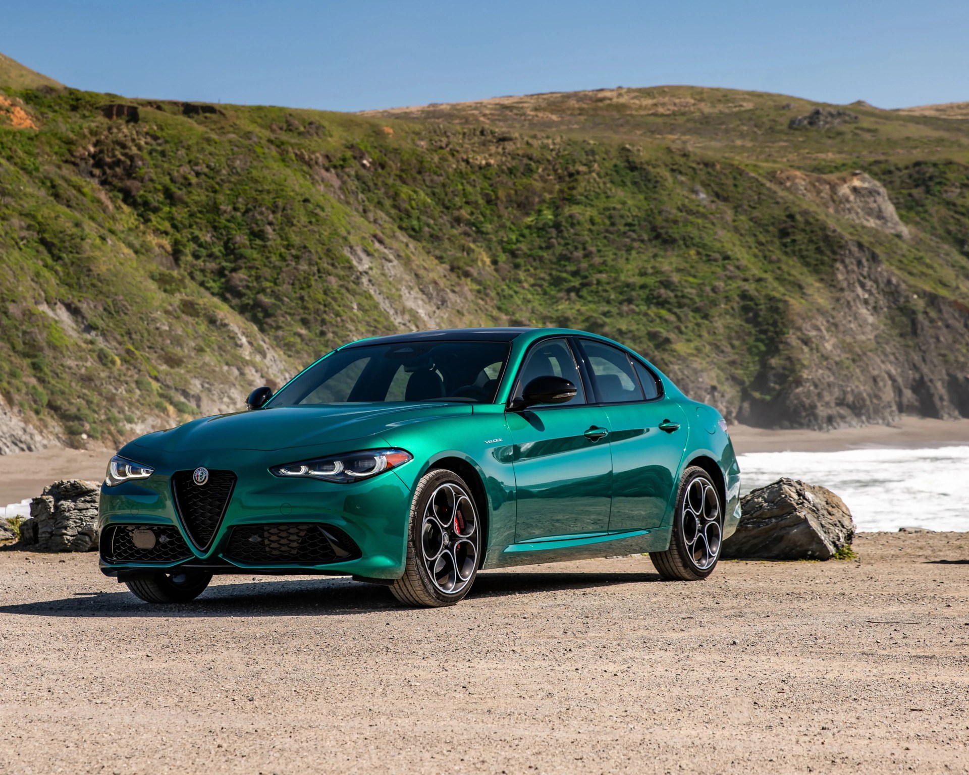 Green Alfa Romeo sedan parked on a sandy beach with rocky hills in the background.
