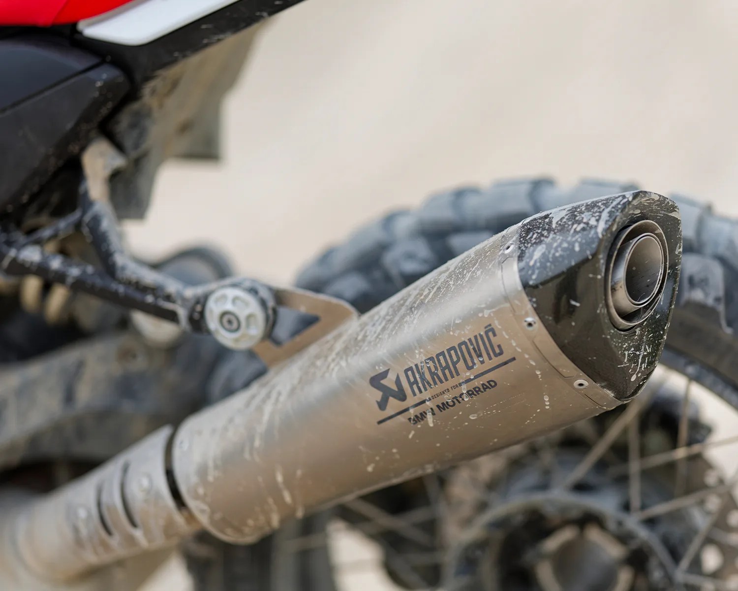 Close-up of a dirty Akrapovič exhaust pipe on a motorcycle with knobby tires in the background.