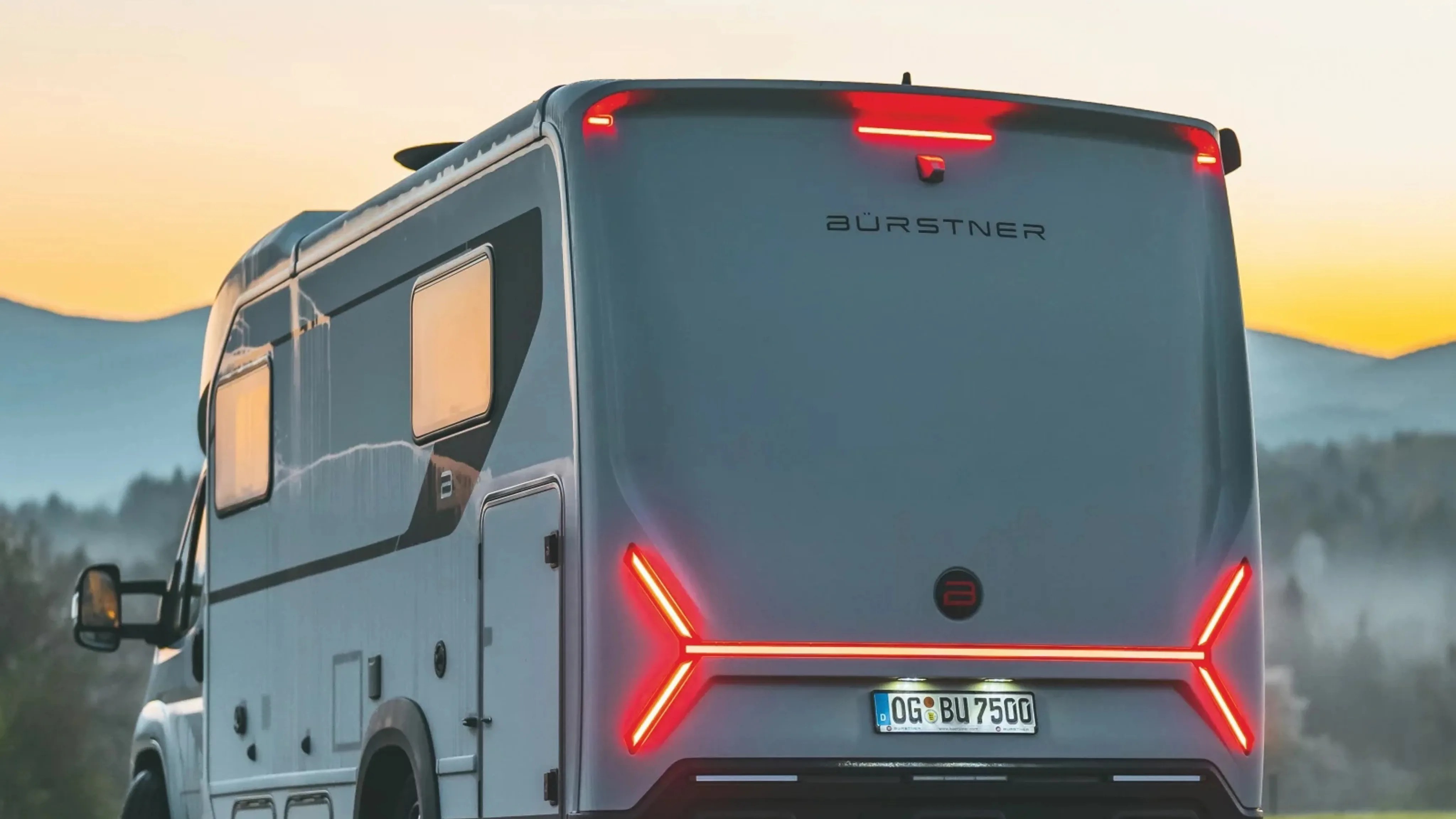 Rear view of a gray Bürstner camper van with illuminated red taillights at sunset.