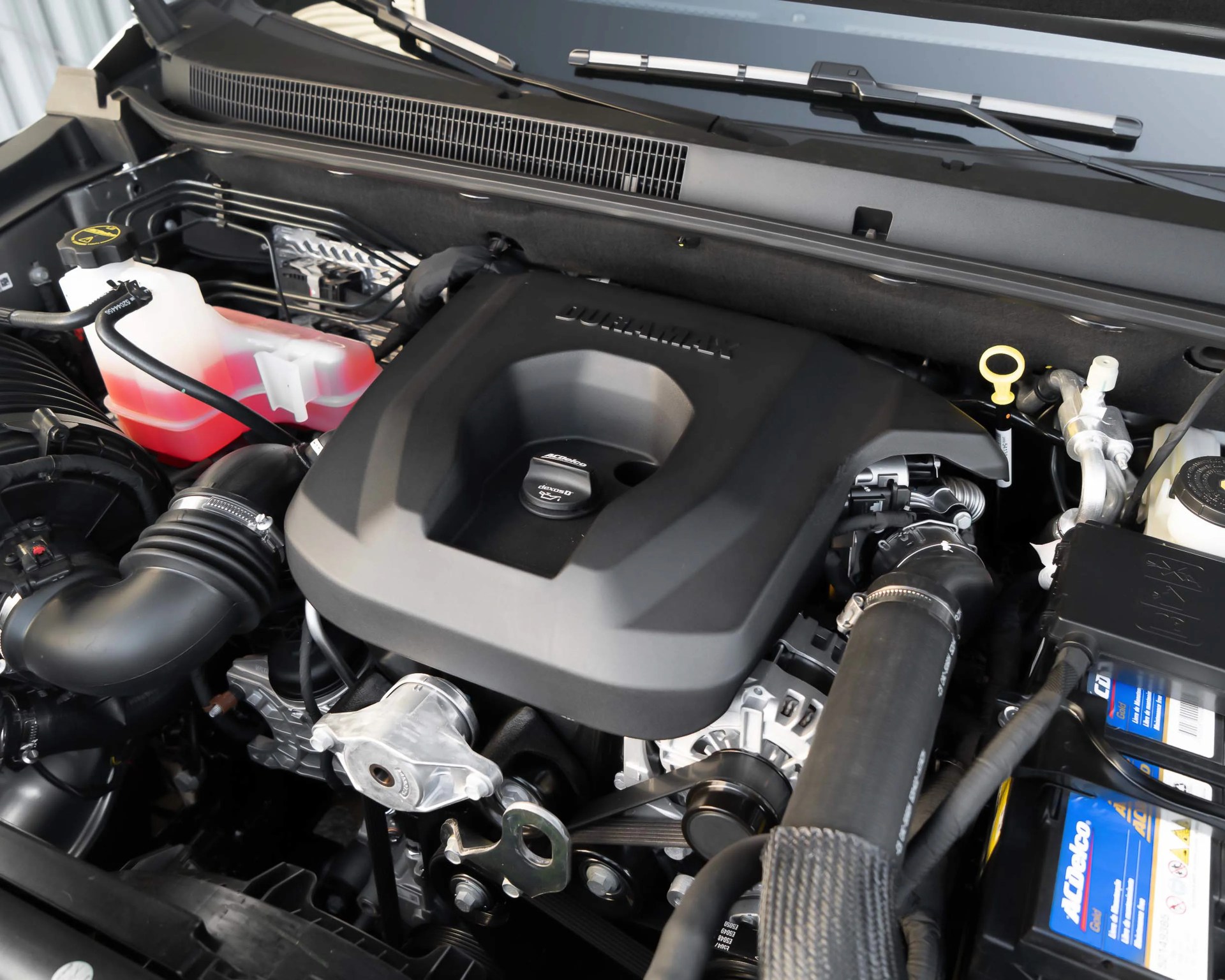 Close-up of a Duramax engine with black plastic cover, coolant reservoir with red fluid, and AC Delco battery.