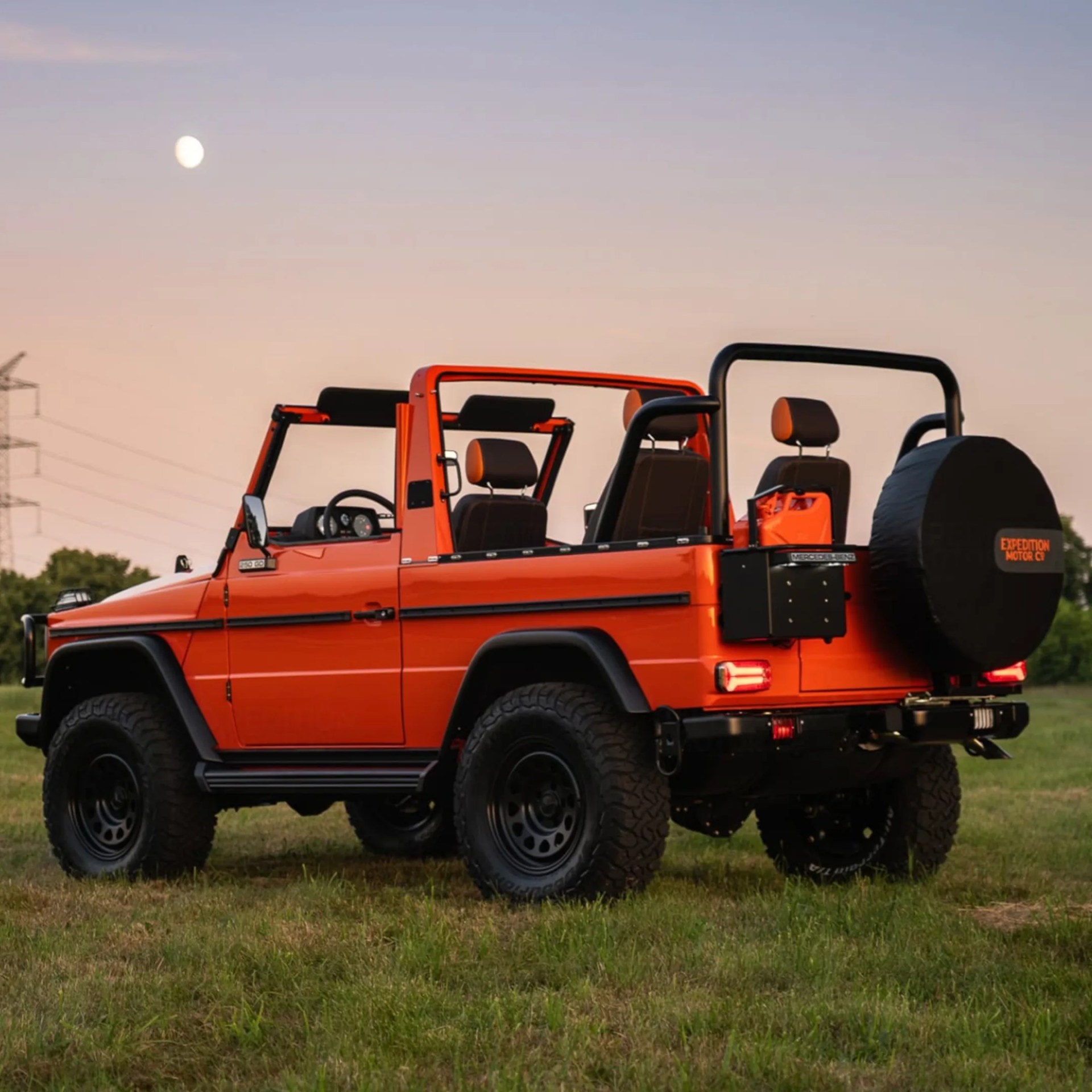 Orange open-top off-road vehicle with black roll bars and spare tire cover, parked on grass at dusk.
