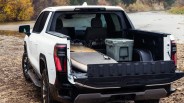 White pickup truck with open tailgate carrying a paddleboard and a gray cooler near a riverbank.