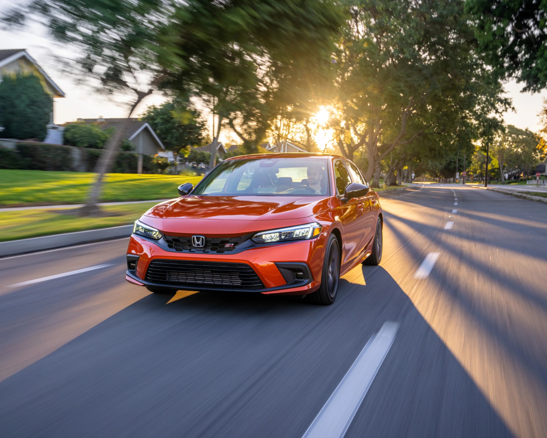 Red Honda Civic Si sedan driving on a sunlit suburban street with trees and houses in the background.