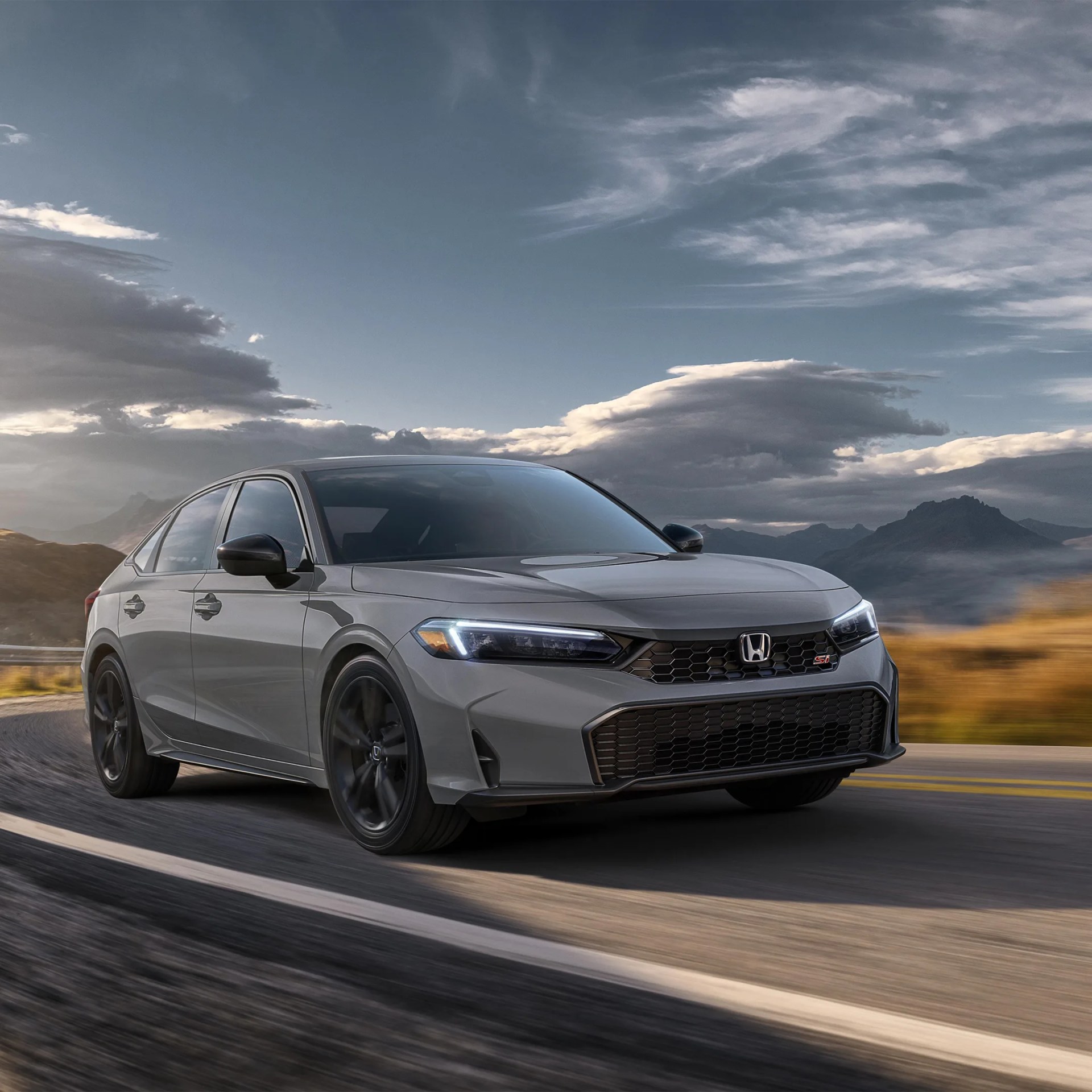 Gray Honda sedan with black wheels driving on a road with mountains and cloudy sky in the background.