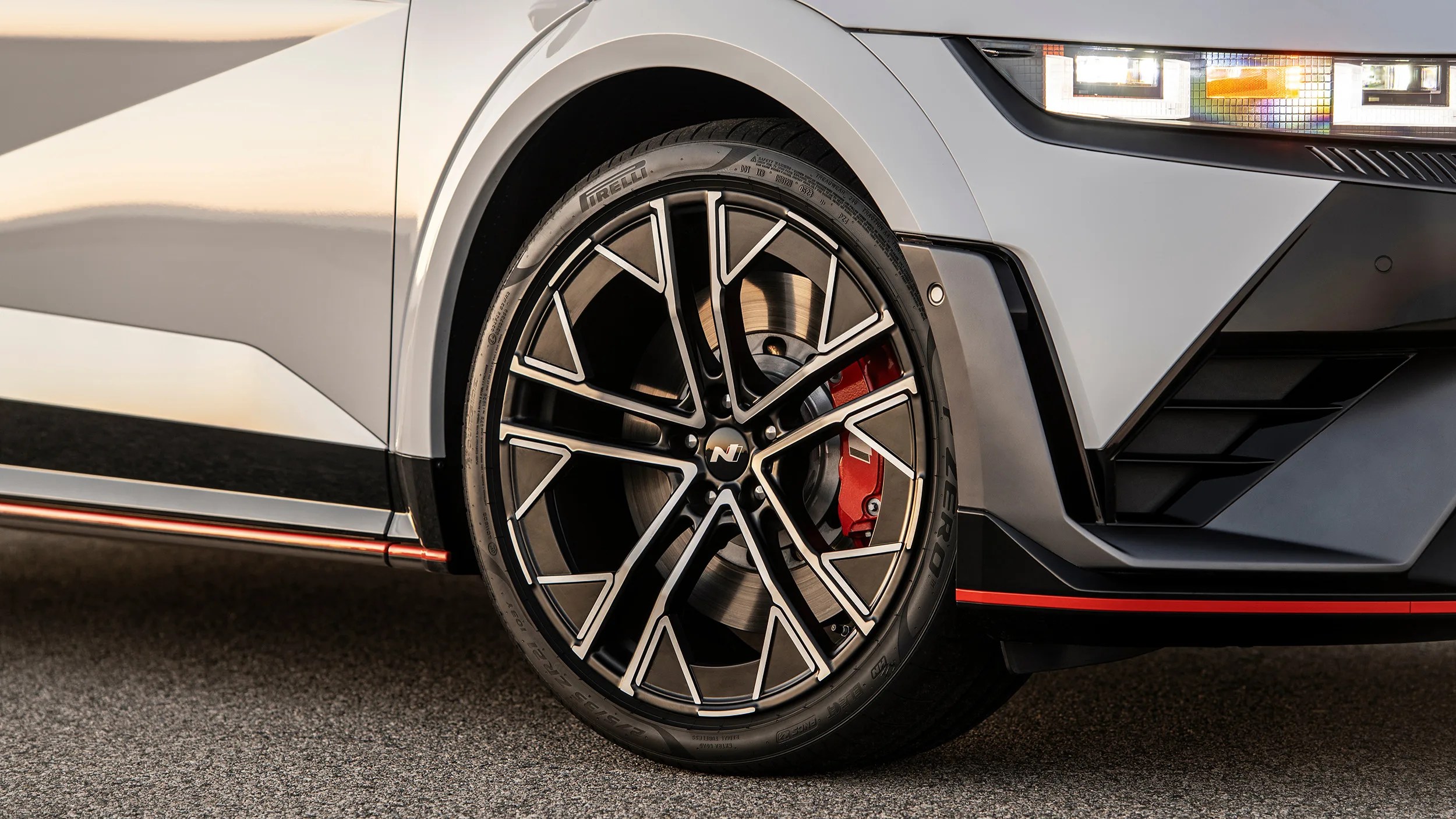 Close-up of a black and silver alloy wheel with red brake caliper on a white car with LED headlight.
