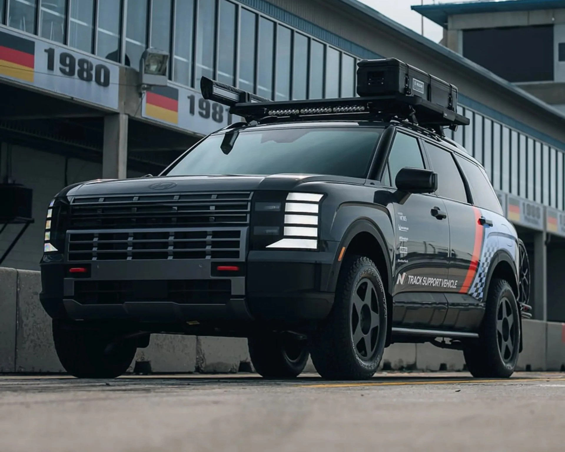 Black SUV with roof rack and storage box parked near a building with large windows and German flag banners.