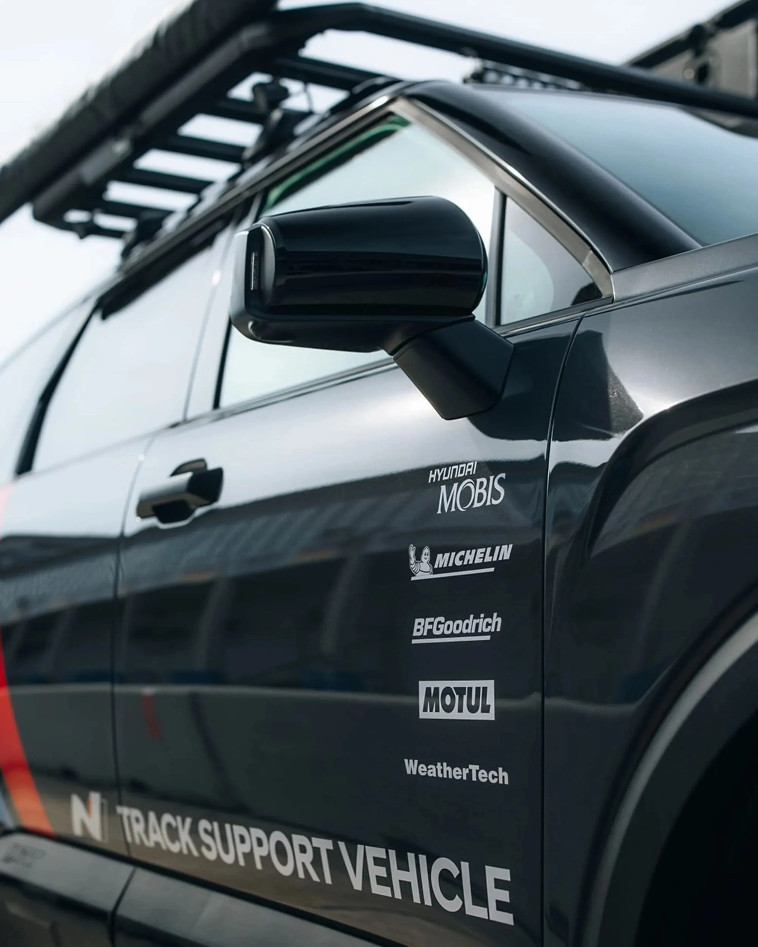 Black side mirror and door of a vehicle with sponsor logos including Hyundai Mobis, Michelin, BFGoodrich, Motul, WeatherTech, and "Track Support Vehicle" text.