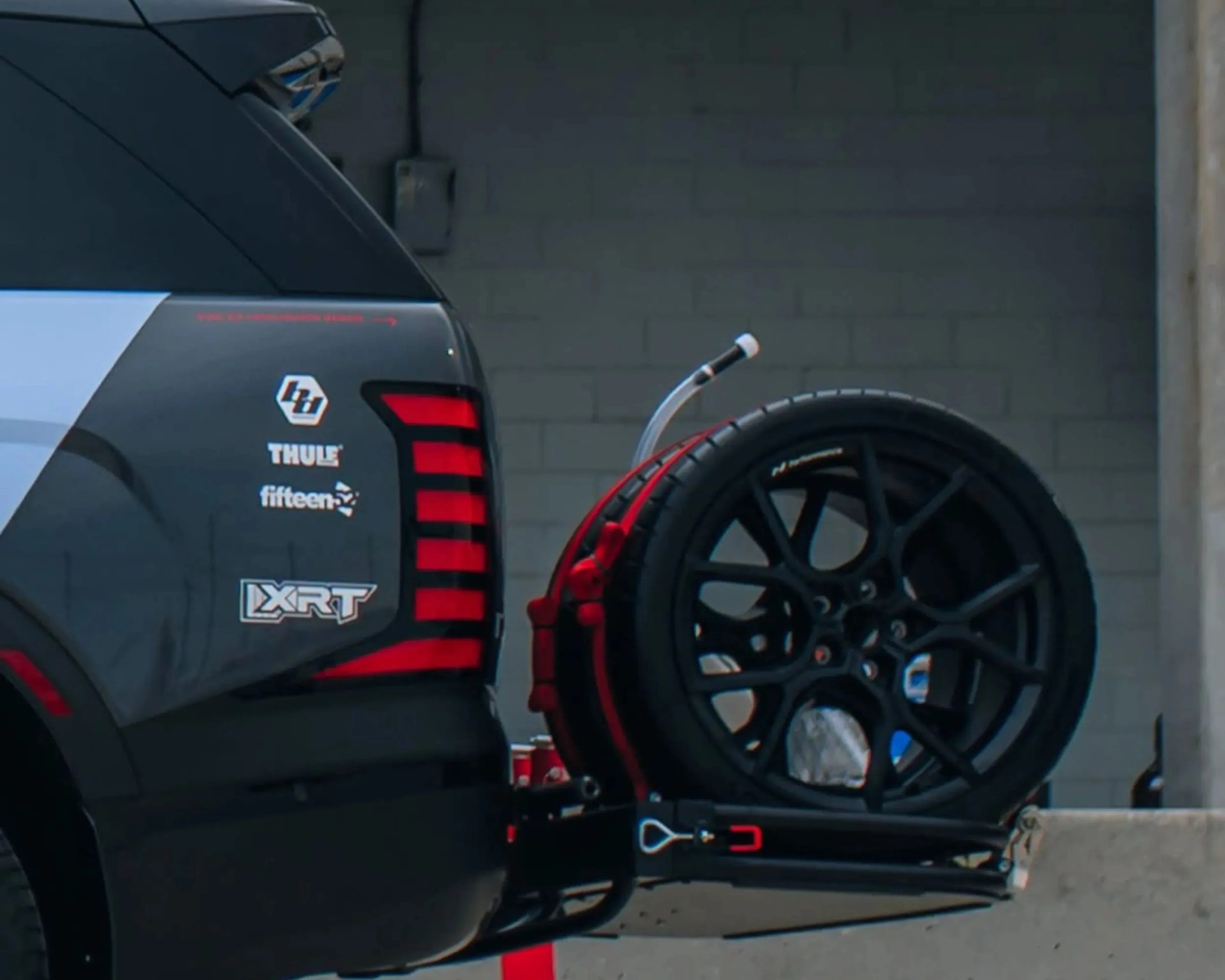 Rear side of a black vehicle with red taillights and logos, carrying a black spare tire mounted on a red carrier.