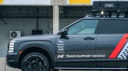 Black SUV with roof cargo box and "Track Support Vehicle" text on the side, parked in front of a concrete wall.