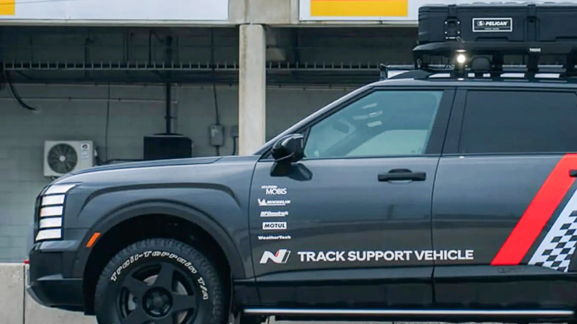 Black SUV with roof cargo box and "Track Support Vehicle" text on the side, parked in front of a concrete wall.