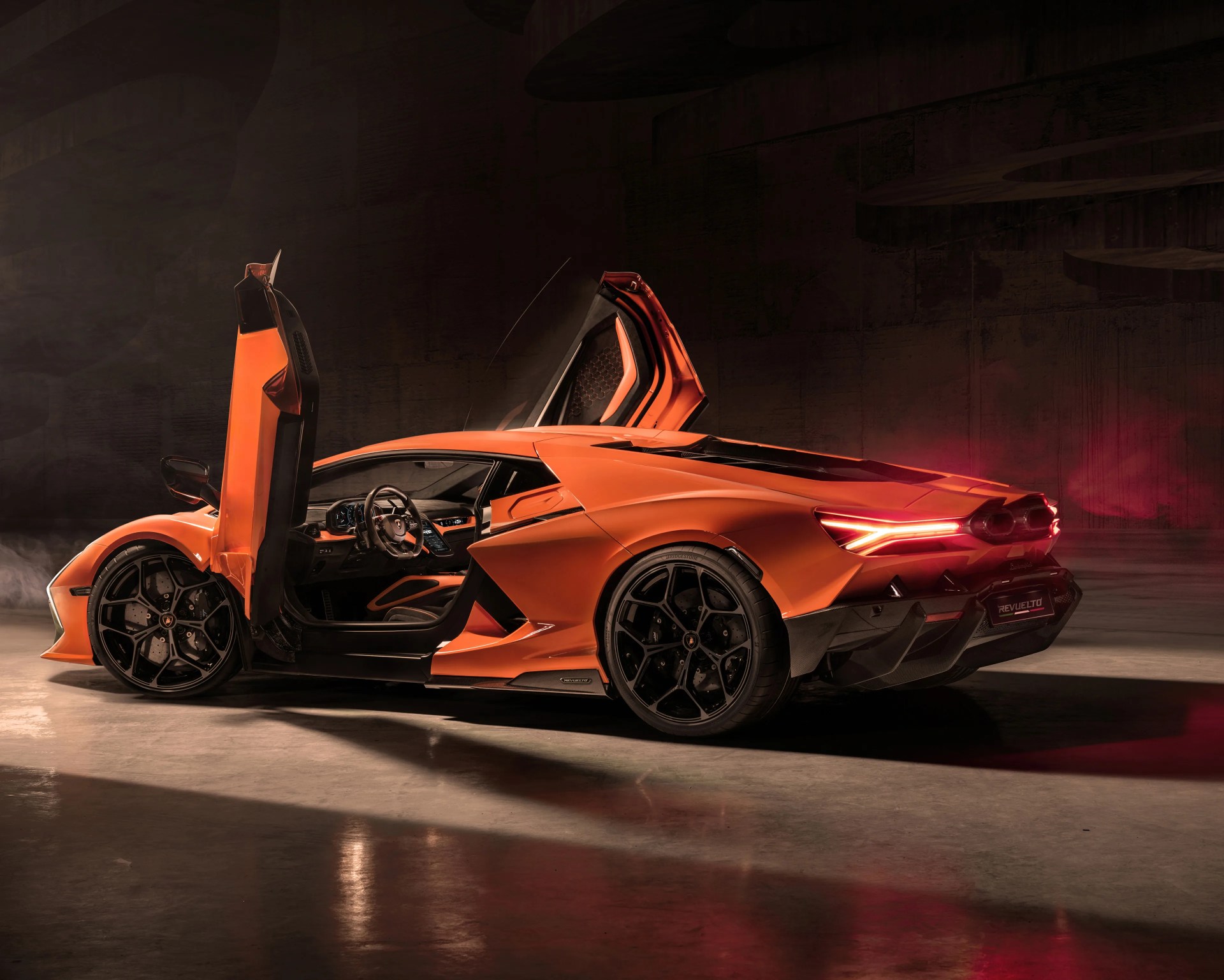 Orange Lamborghini sports car with scissor doors open, black wheels, and illuminated rear lights in a dark setting.