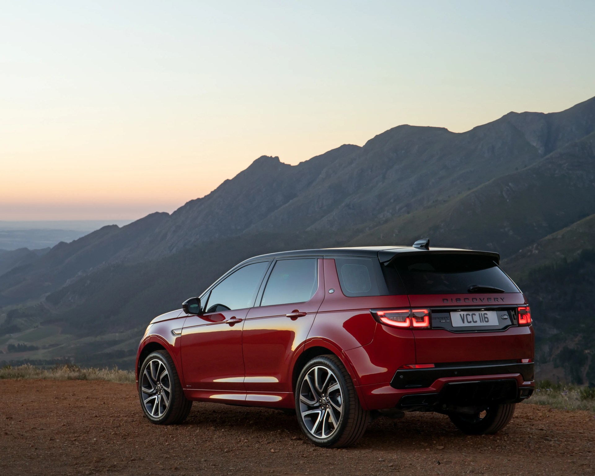Red Land Rover Discovery Sport with black roof parked on dirt road with mountain backdrop at sunset.