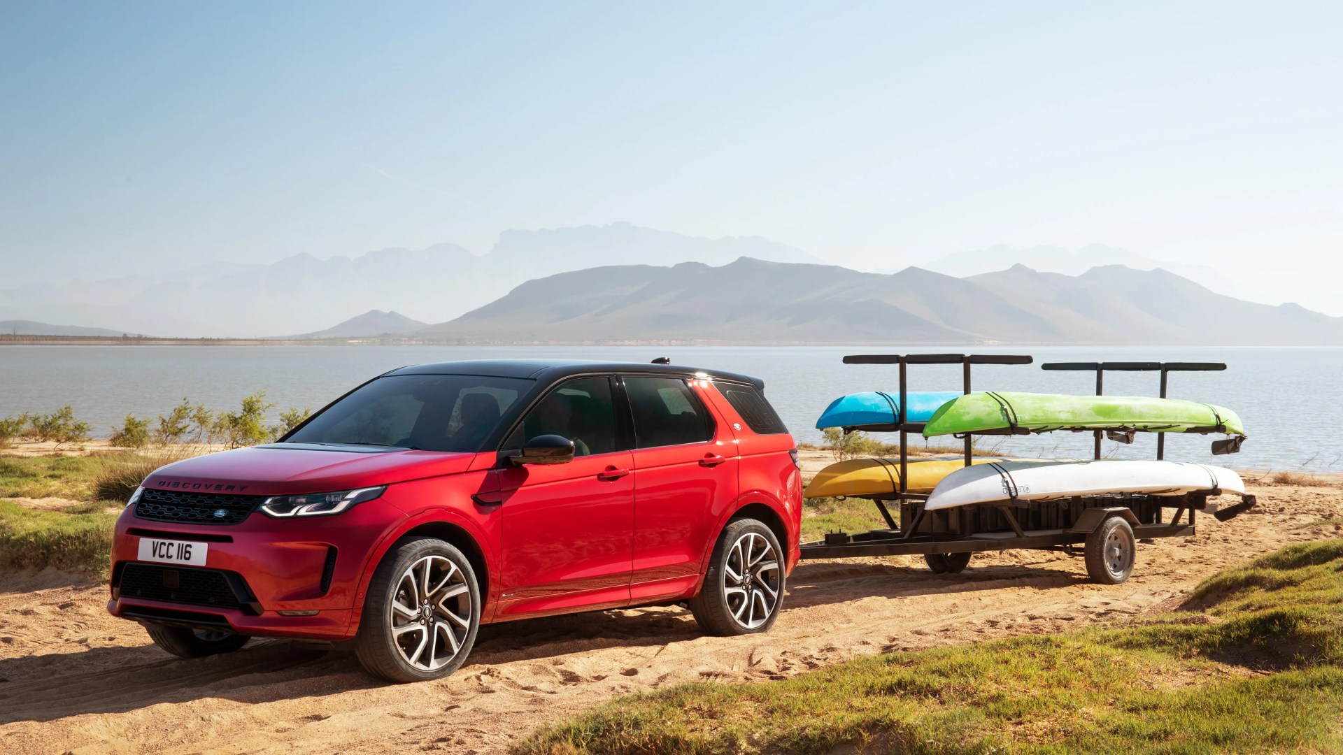 Red Land Rover Discovery SUV with black roof towing a trailer carrying three kayaks on a sandy shore by a lake.