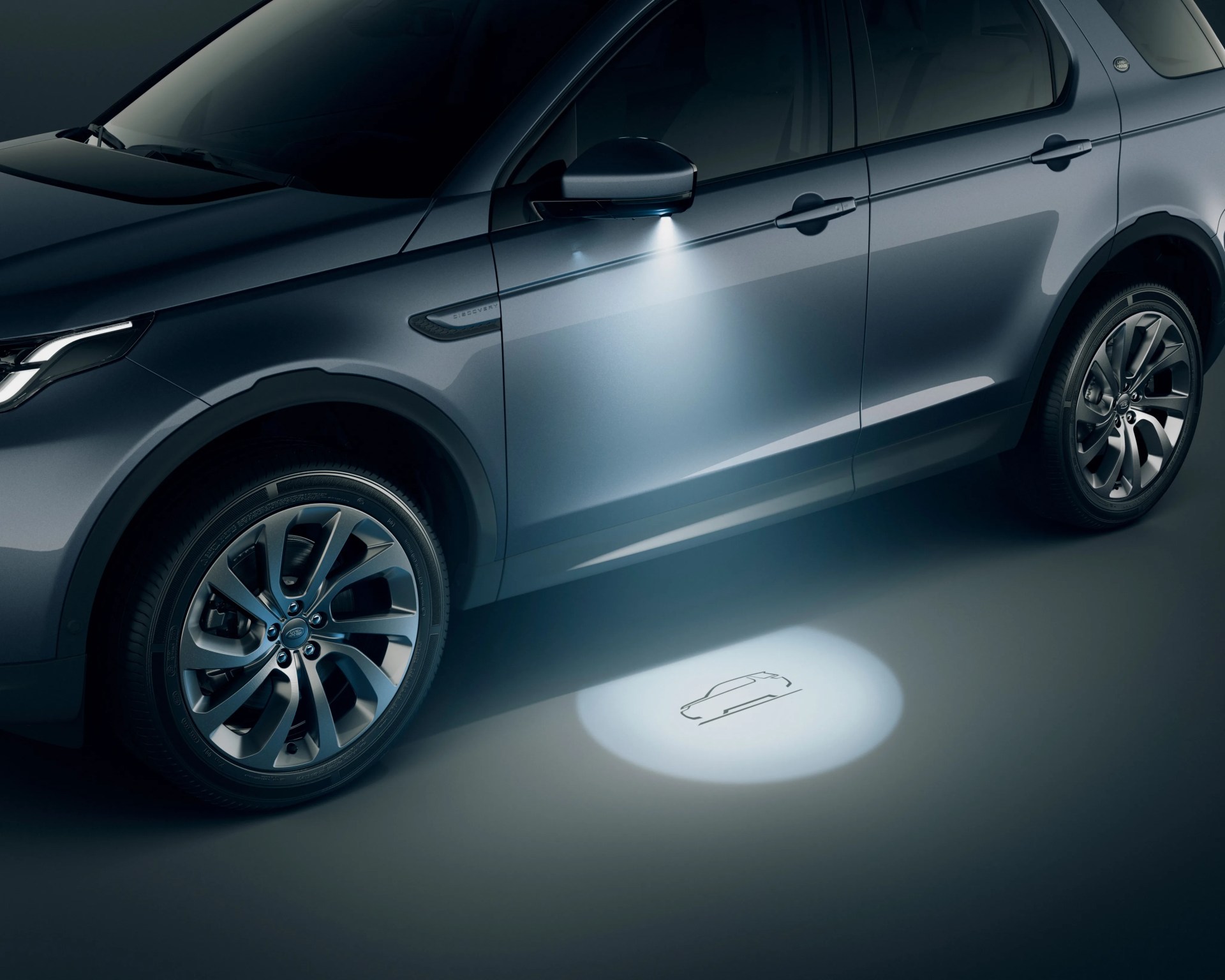 Blue Land Rover Discovery with illuminated side mirror projecting car outline on the ground.