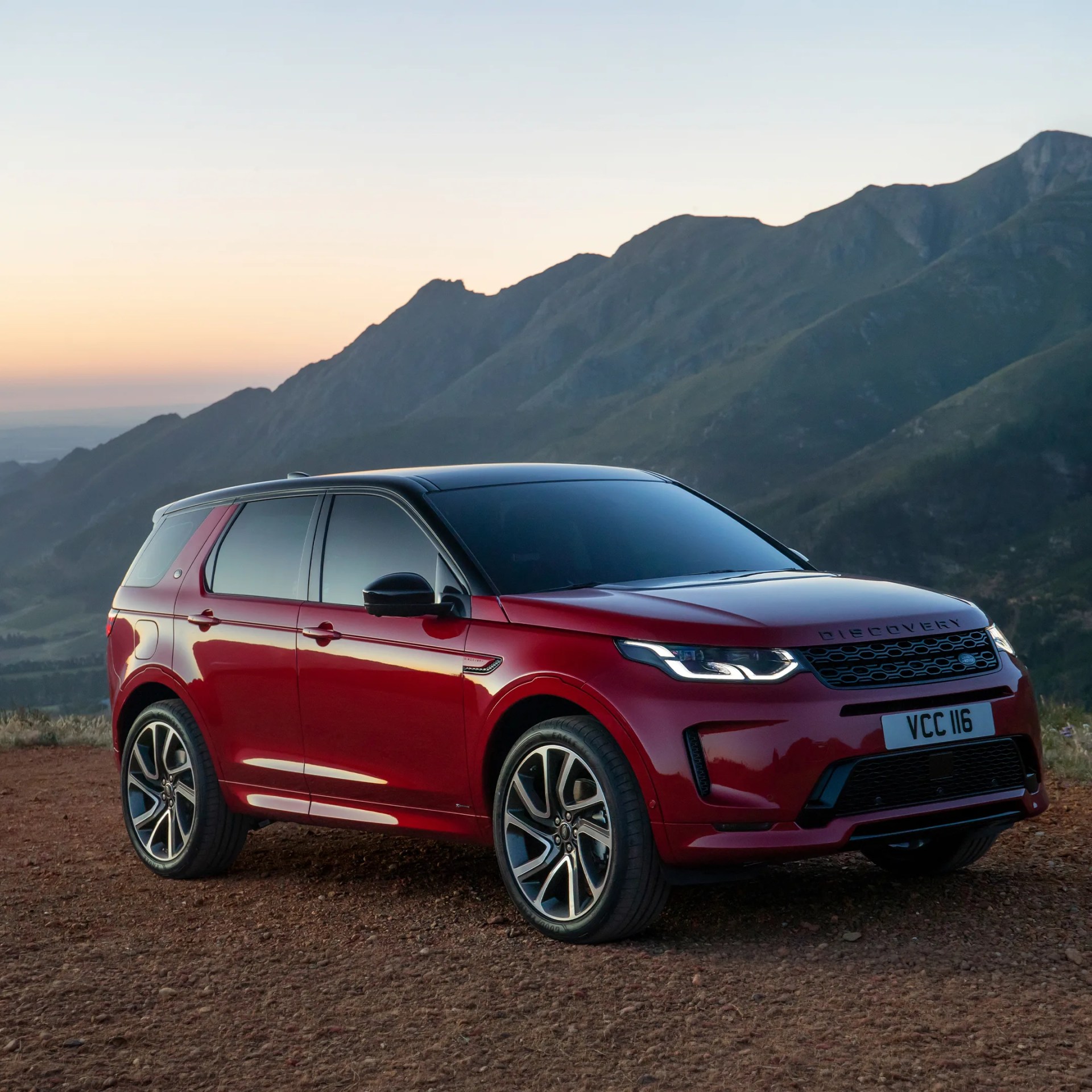 Red Land Rover Discovery SUV with black roof and alloy wheels parked on dirt with mountains in background at sunset.