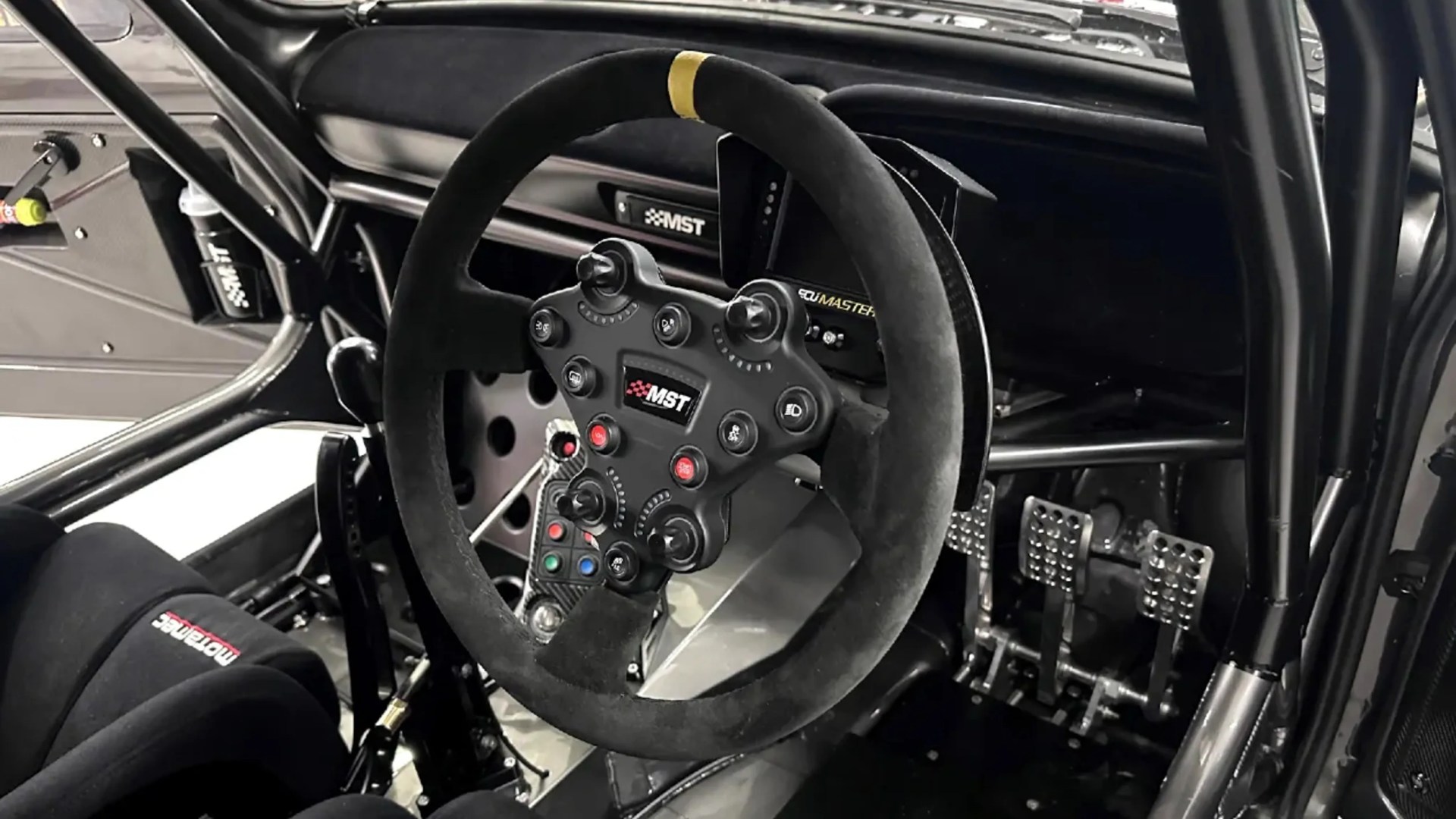 Black suede racing steering wheel with multiple buttons and MST logo inside a race car cockpit.