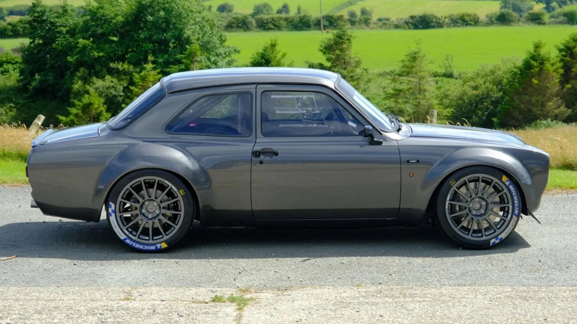 Gray classic two-door car with wide tires and Michelin branding parked on a road with green fields in the background.