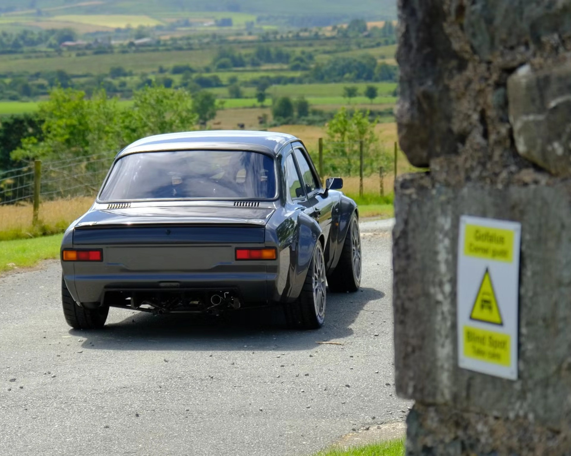 Dark gray classic car with wide tires parked on a rural road near a stone wall with warning signs.
