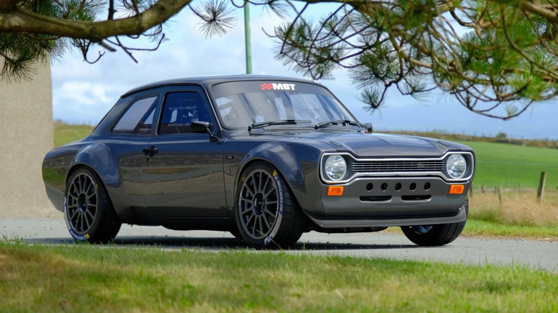 Dark gray vintage rally car with wide fenders and racing tires parked on a road near grass and trees.
