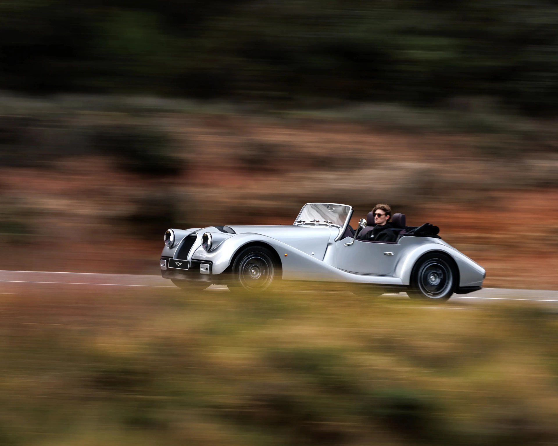 Silver classic convertible car driving on a road with blurred natural background.