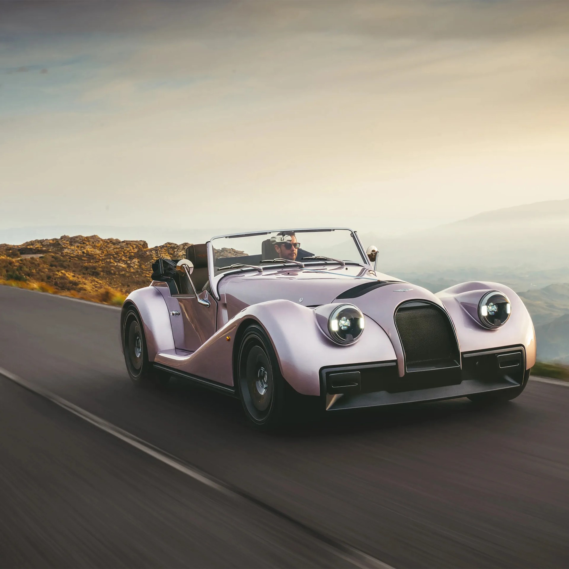 Lavender vintage convertible car with round headlights driving on a road near rocky hills at sunset.