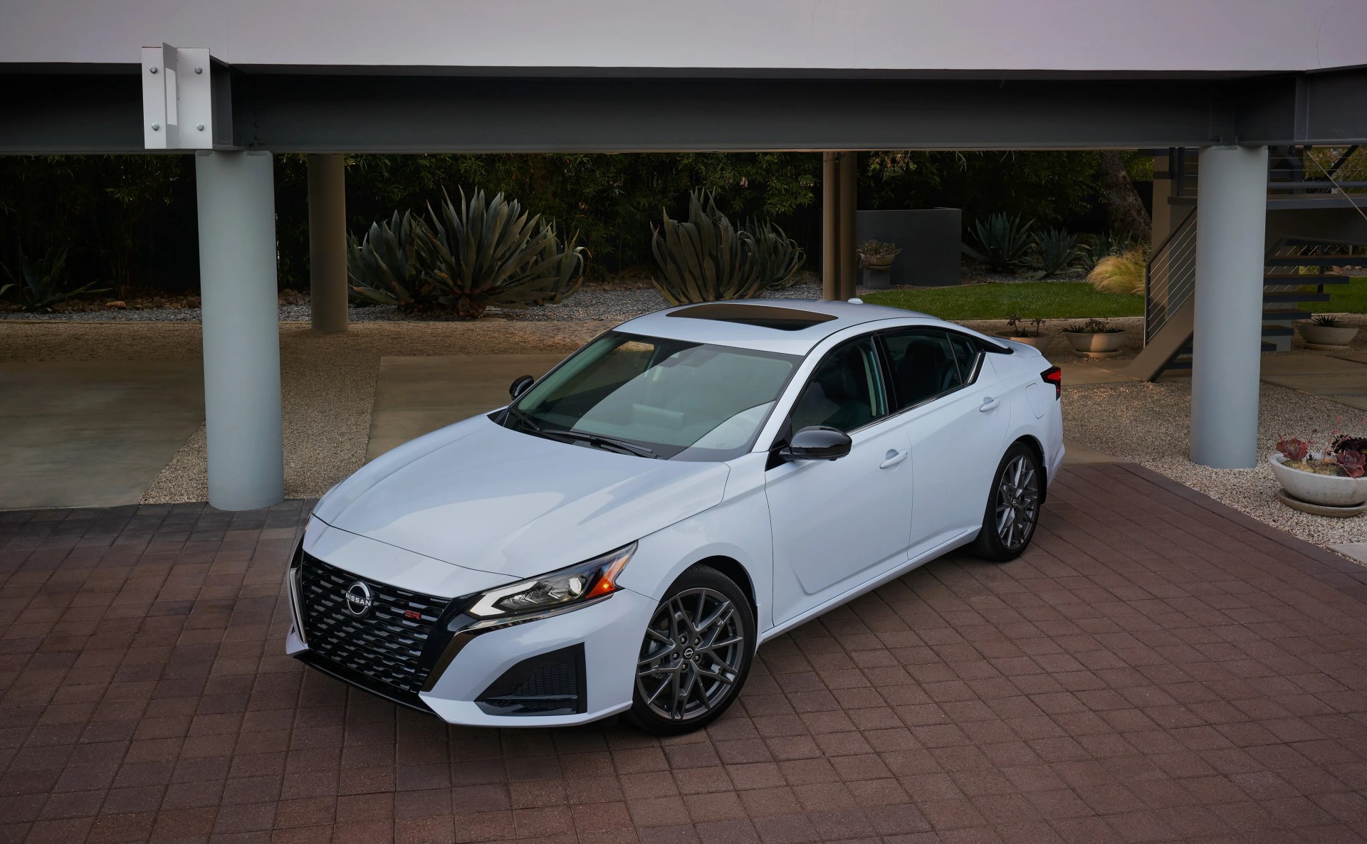 White Nissan sedan with black grille and sunroof parked on a brick driveway under a modern carport.