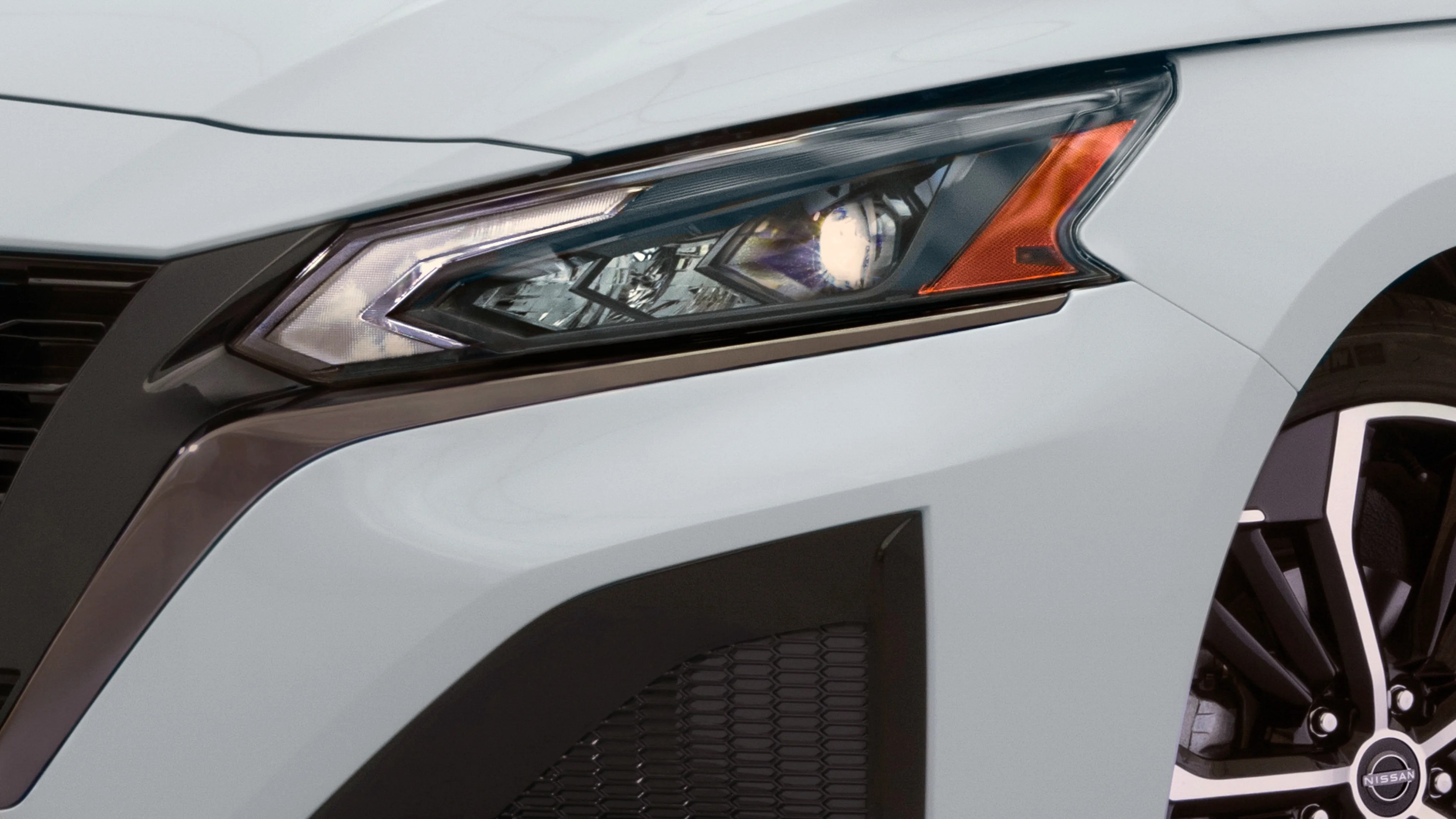 Close-up of a white car's angular headlight with an orange indicator and part of a black and silver Nissan wheel.