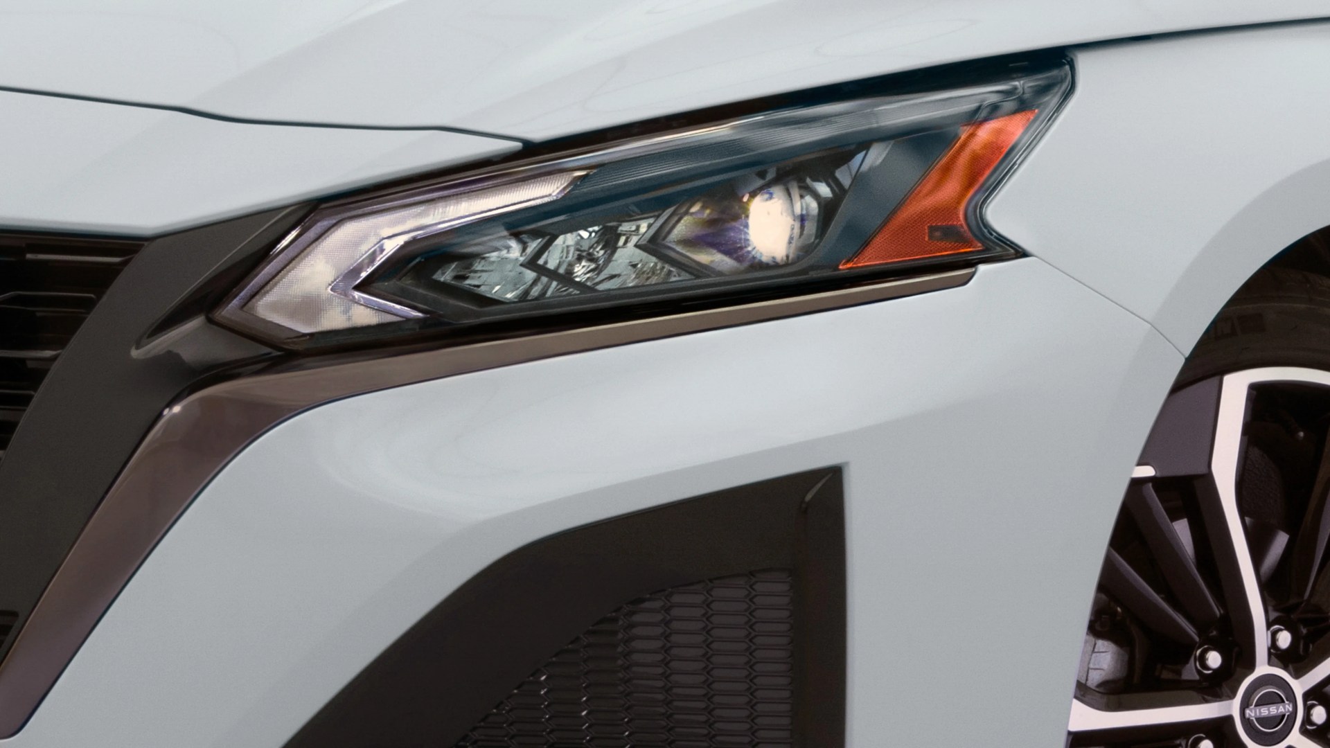Close-up of a white car's angular headlight with an orange indicator and part of a black and silver Nissan wheel.