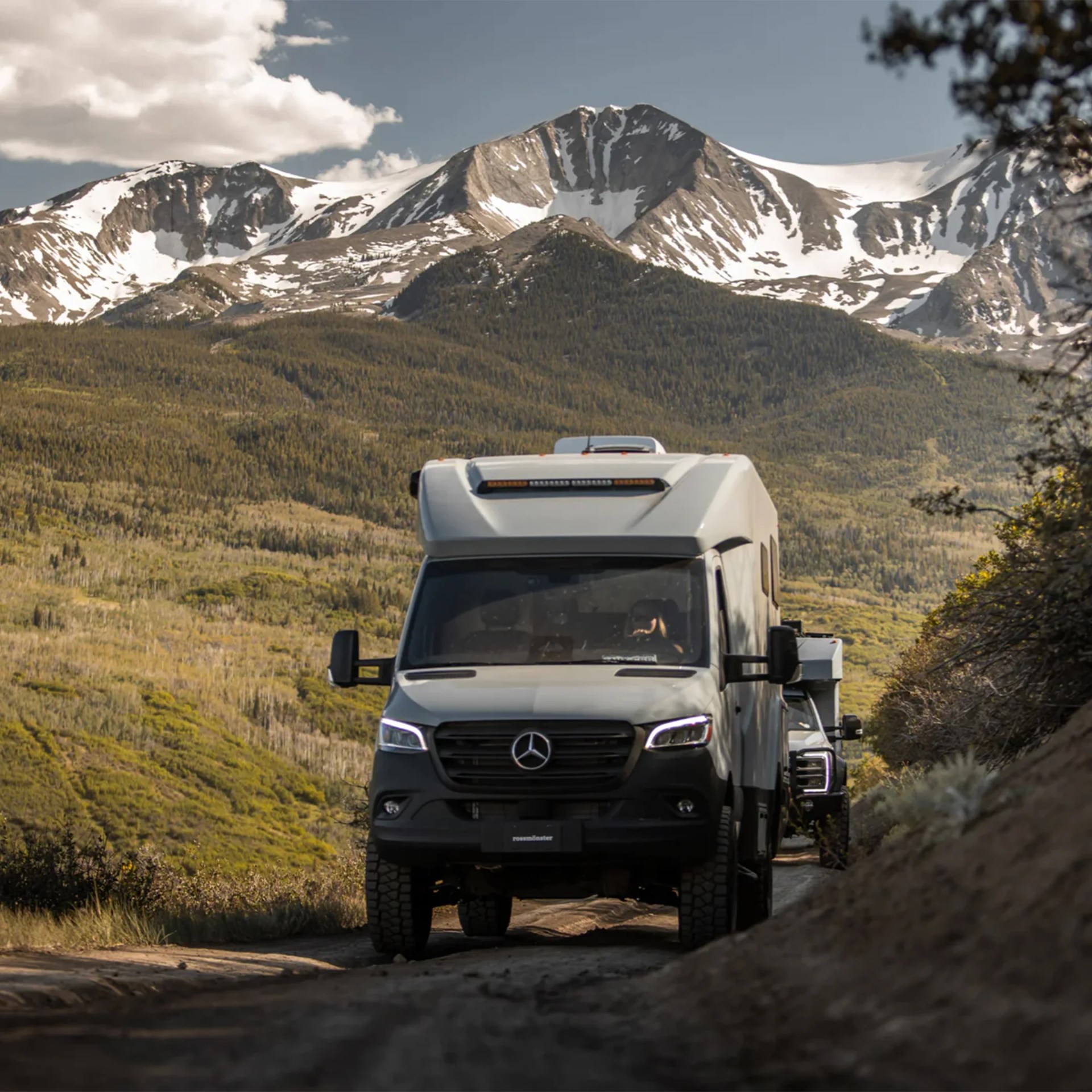White Mercedes-Benz camper van driving on a dirt road with snow-capped mountains in the background.