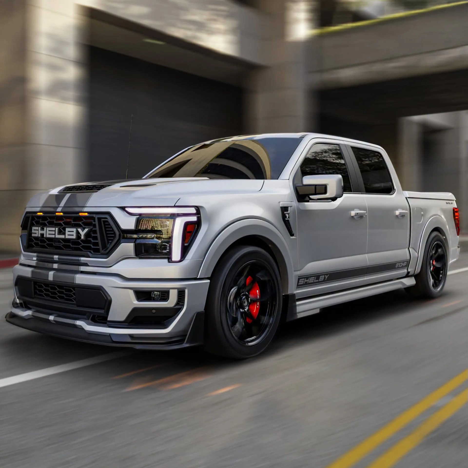Silver Shelby pickup truck with black racing stripes and red brake calipers driving on a city street.