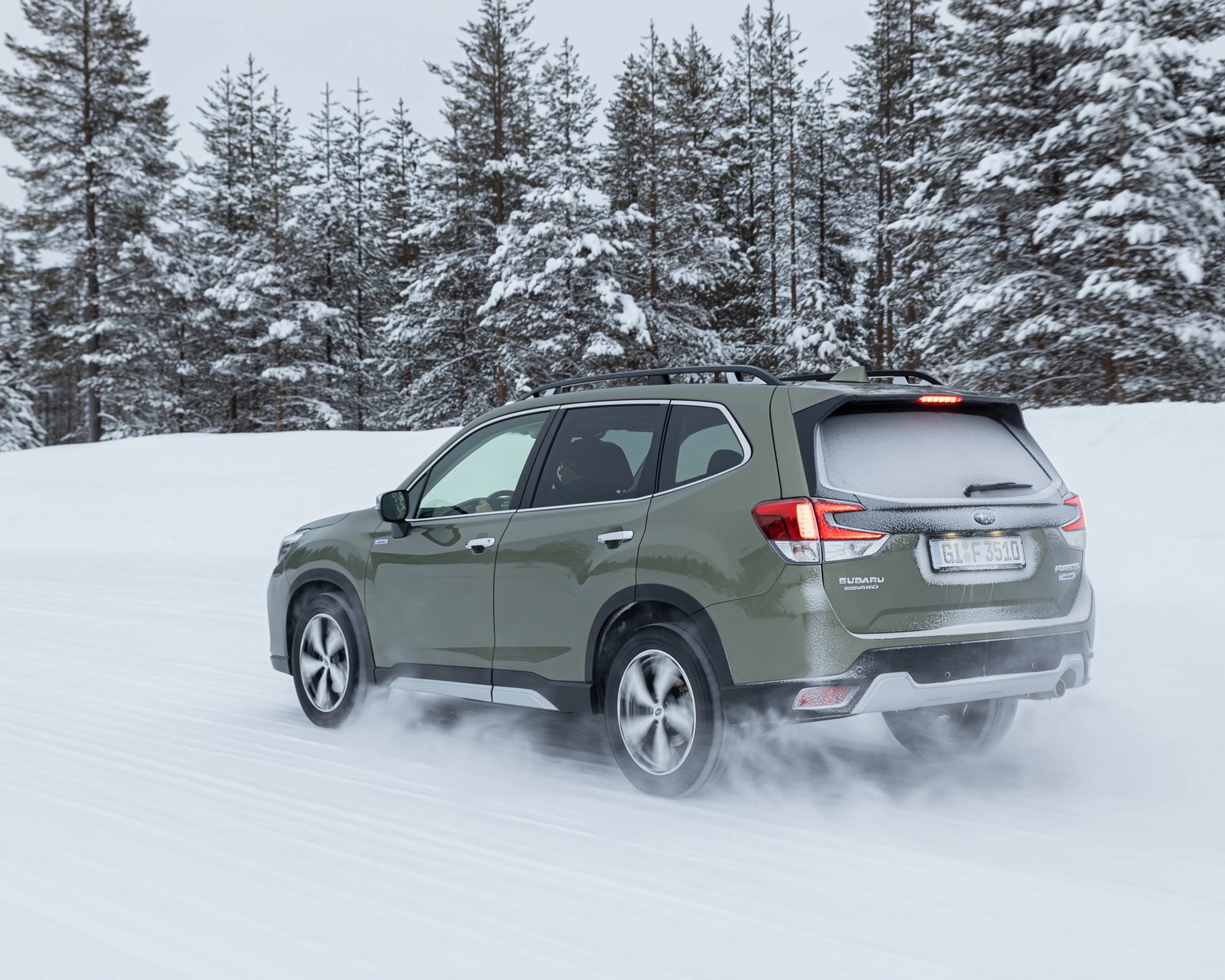Green Subaru Forester AWD SUV driving on snow-covered road with snow-covered pine trees in background.