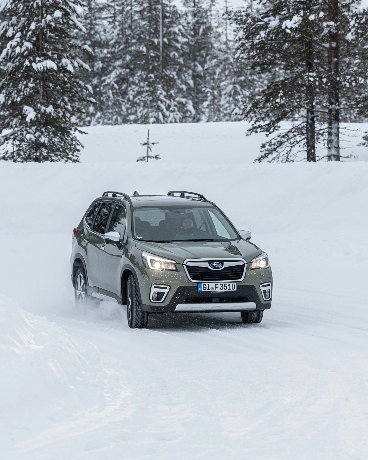 Green Subaru SUV driving on a snowy road with snow-covered trees in the background.