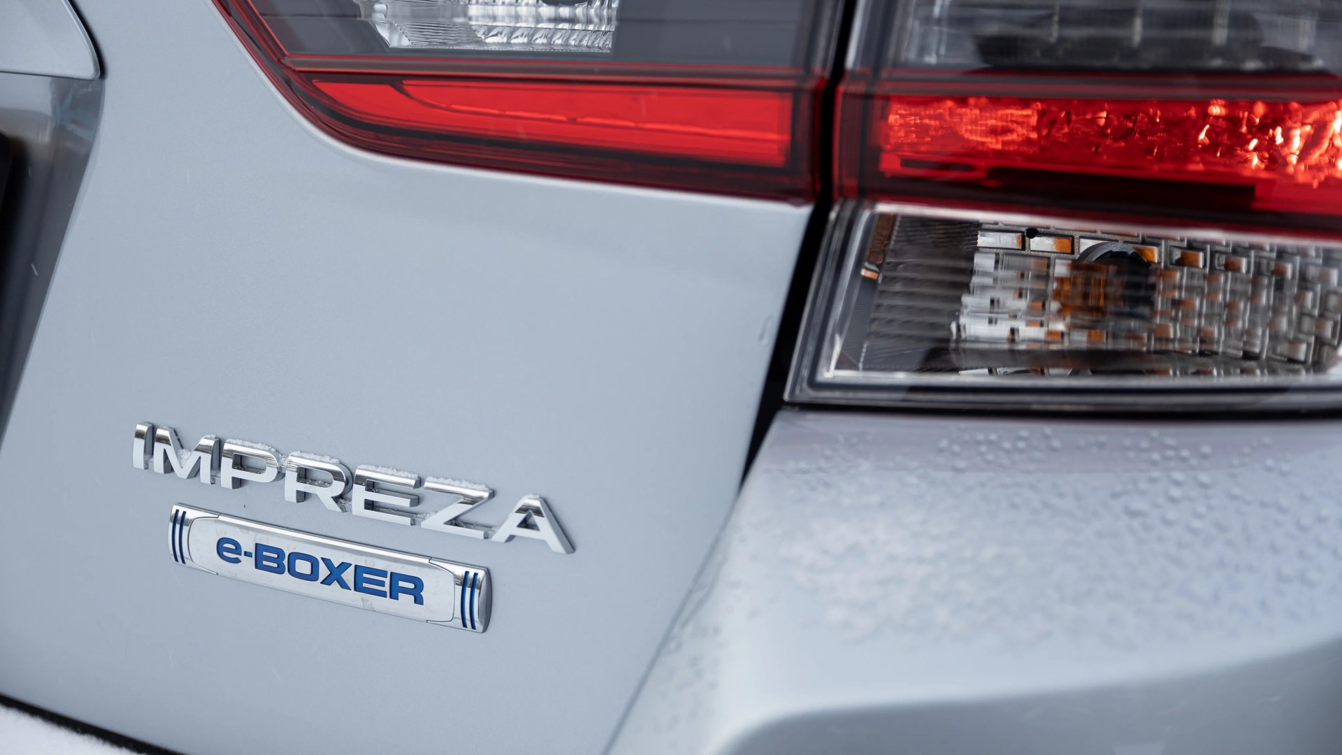 Close-up of a silver Subaru Impreza e-Boxer badge next to a red and clear taillight with water droplets on the car surface.