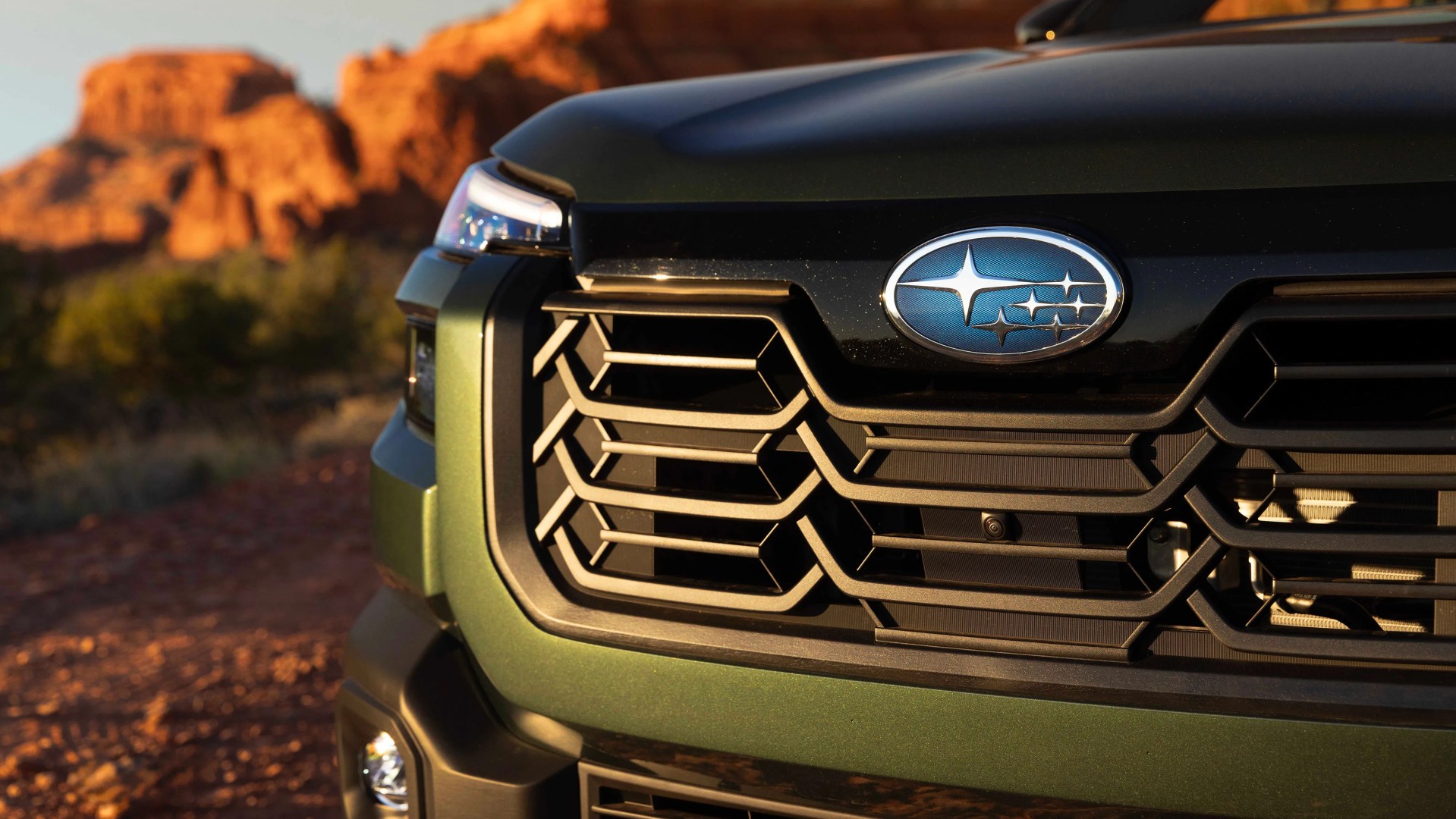 Front grille of a green Subaru vehicle with a blue Subaru logo and a rocky desert background.