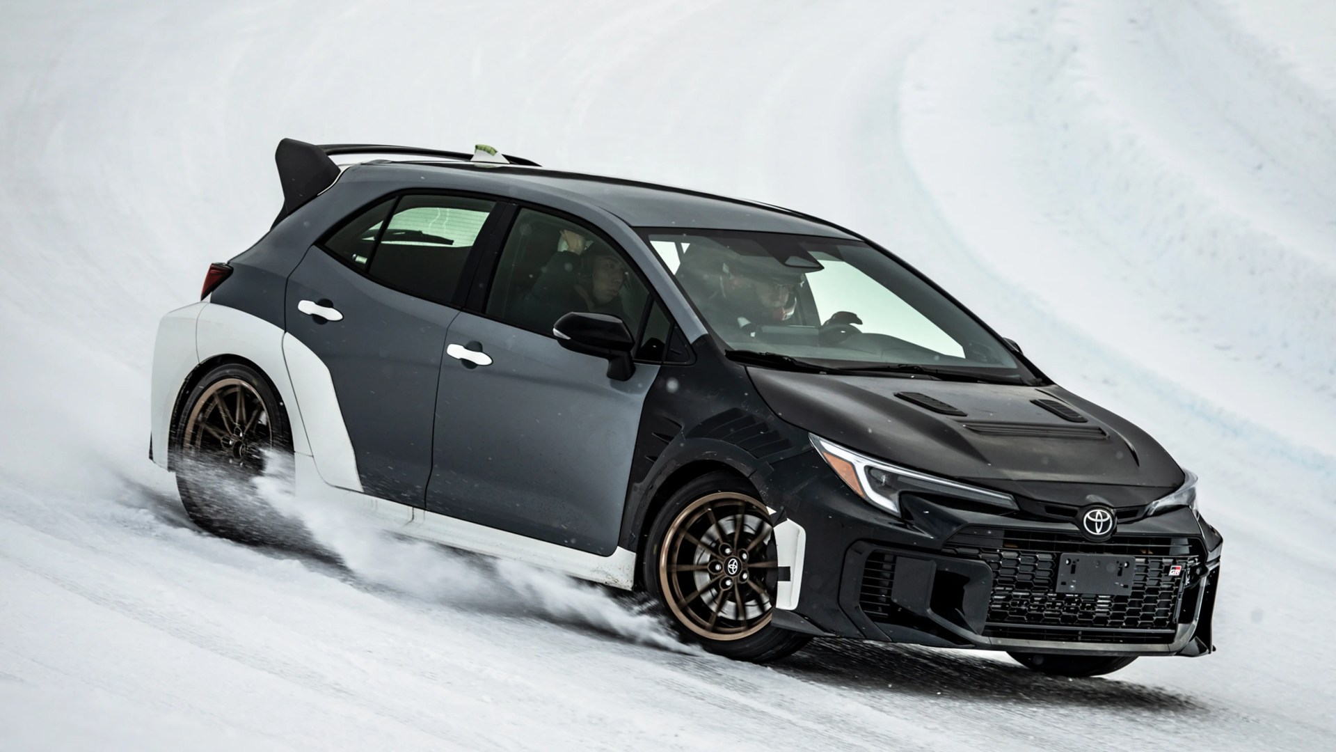 Black and white Toyota GR hatchback drifting on snow with bronze wheels and two people inside.