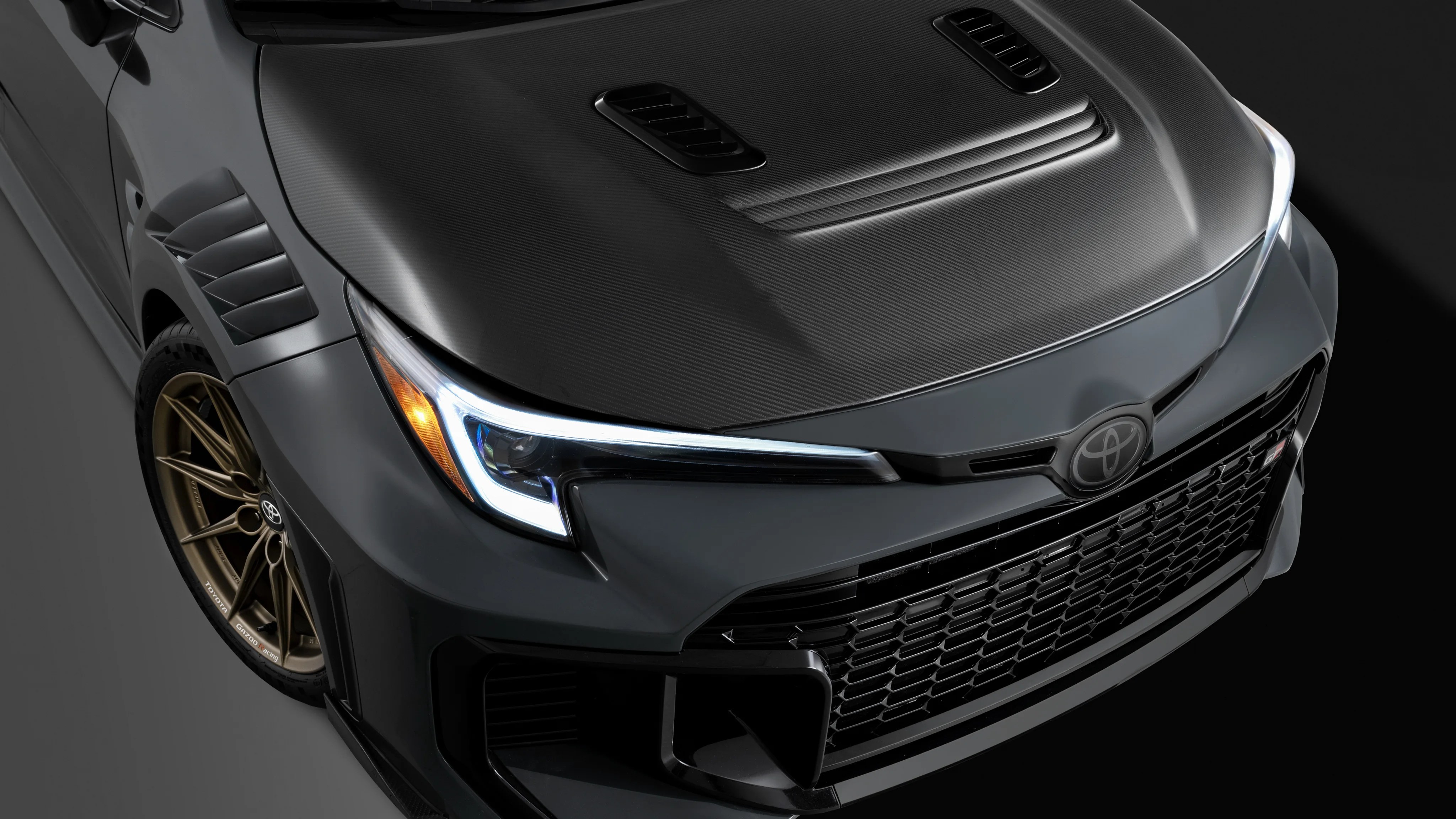 Close-up of a dark gray Toyota car front with carbon fiber hood, bronze alloy wheel, and LED headlight on a black background.