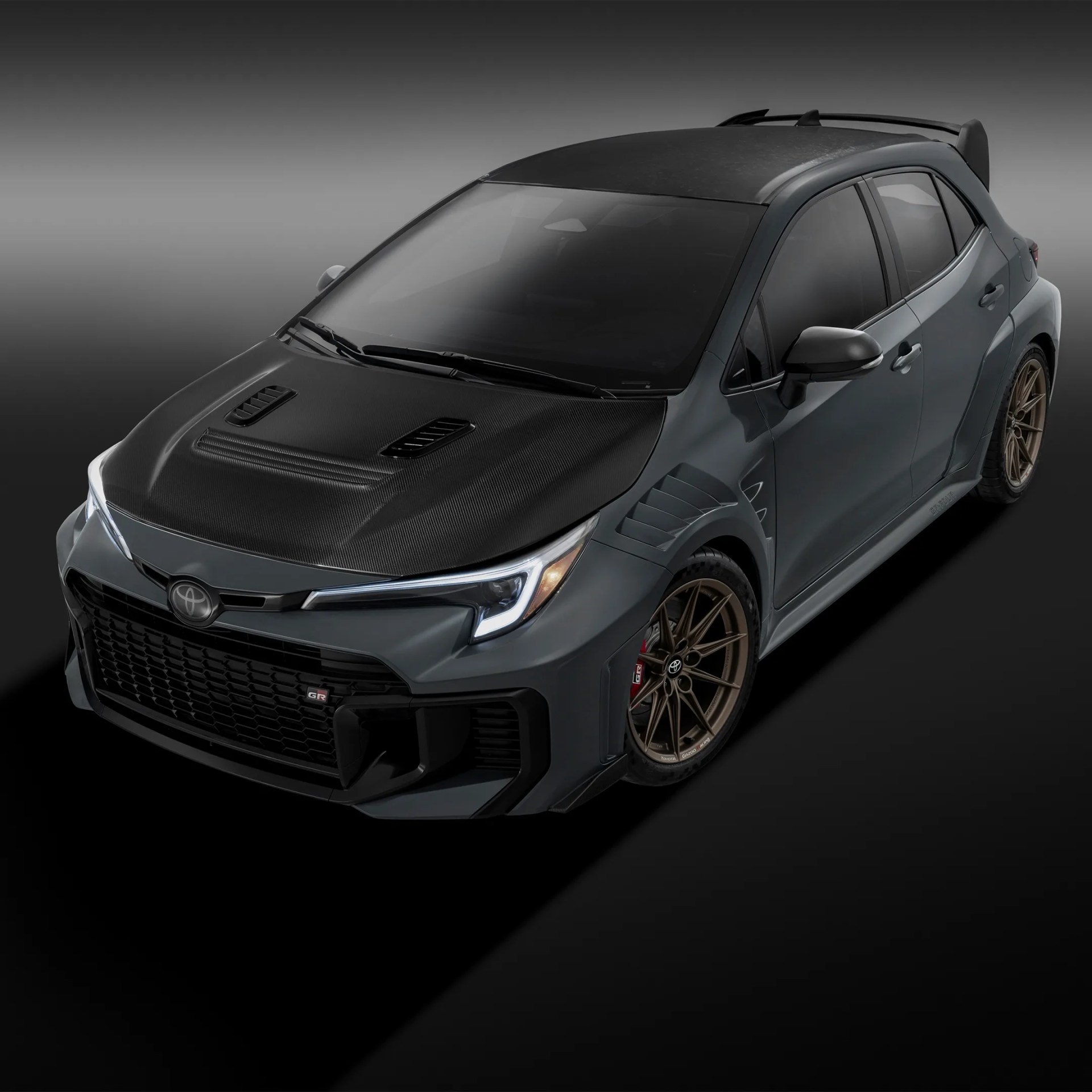 Gray Toyota GR Yaris with carbon fiber hood, bronze wheels, and rear spoiler on a dark gradient background.