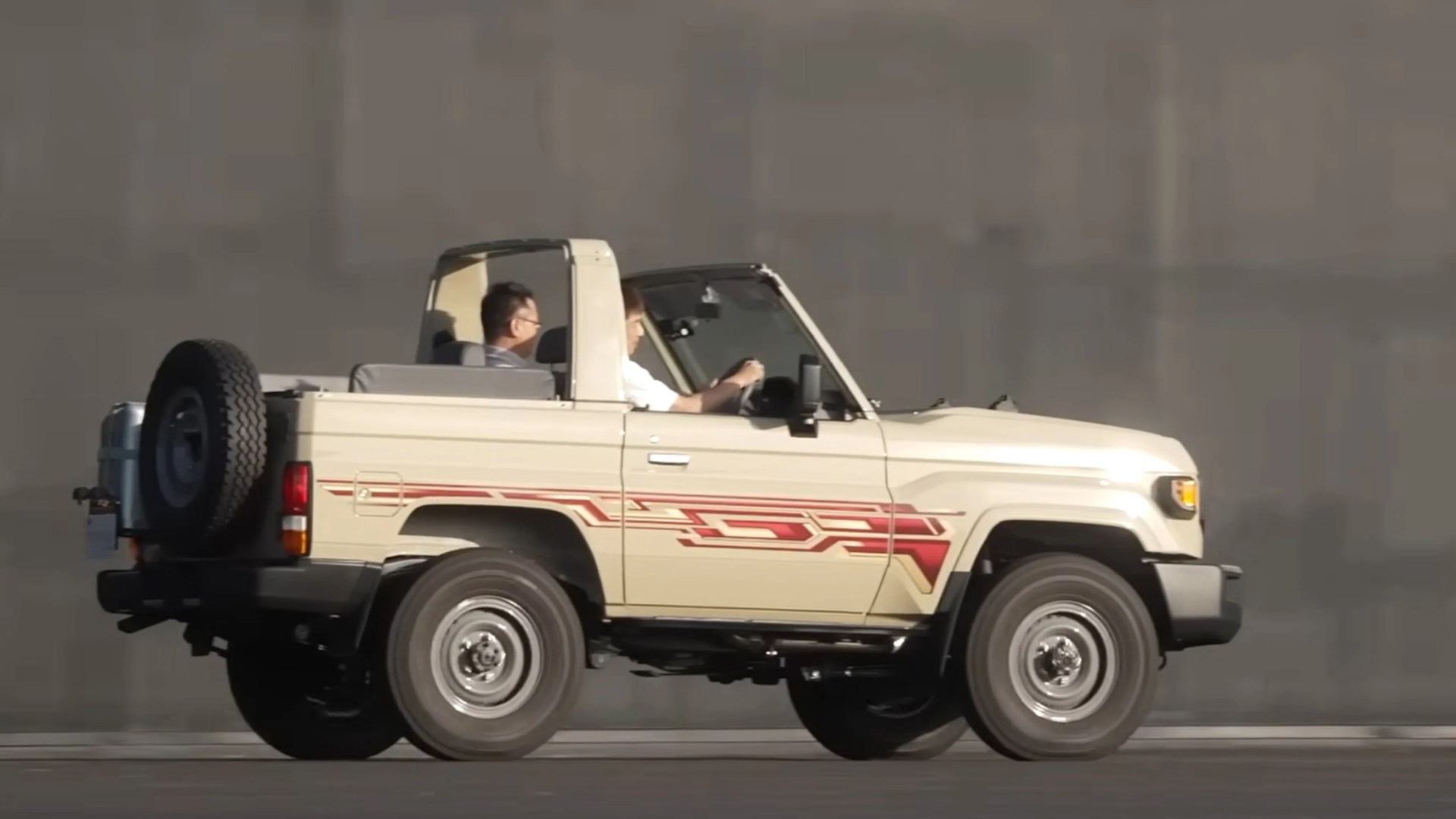 Beige open-top off-road vehicle with red side decals and two passengers inside.