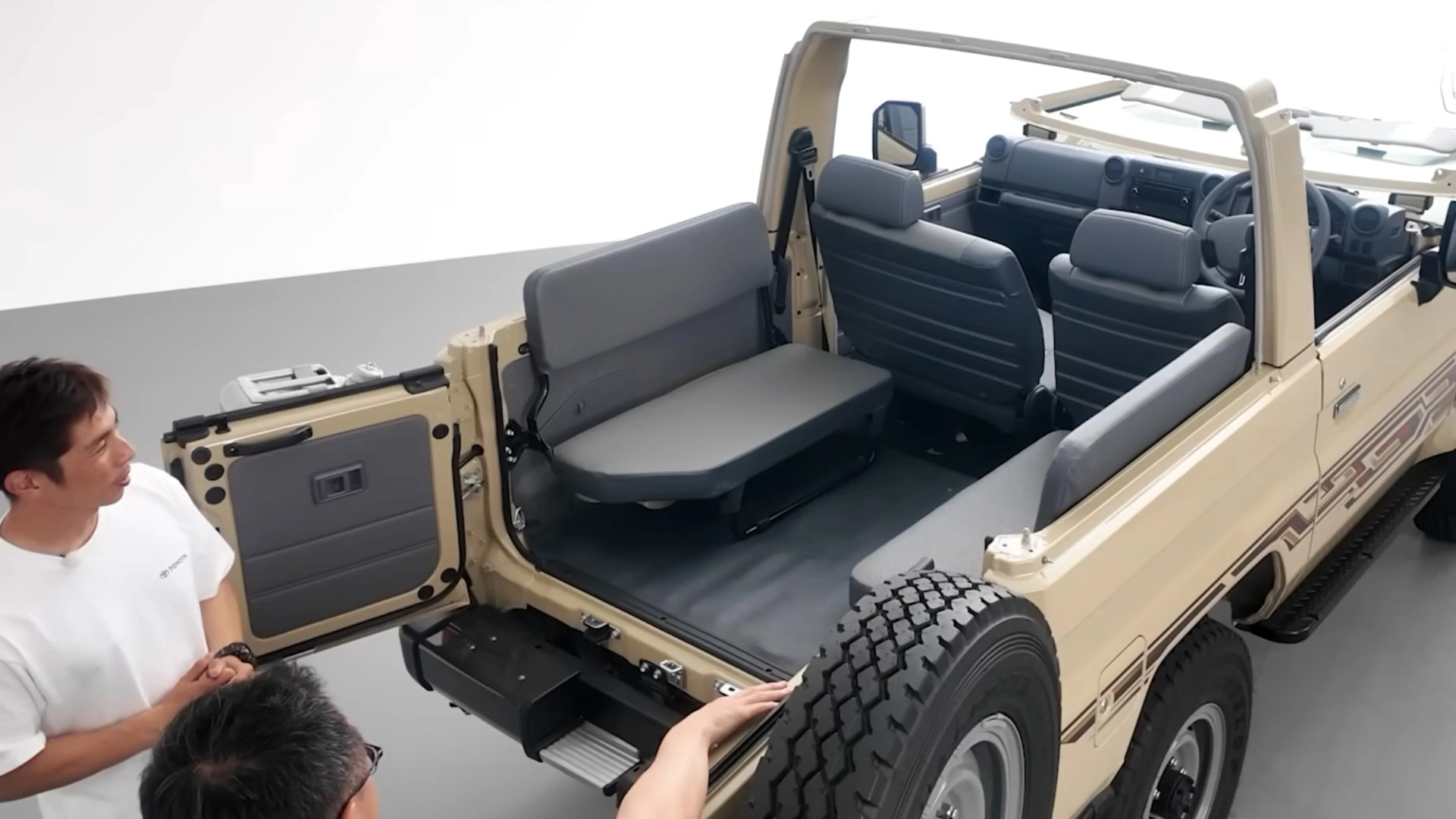 Beige open-top 6x6 off-road vehicle with gray bench seats and two men standing nearby.